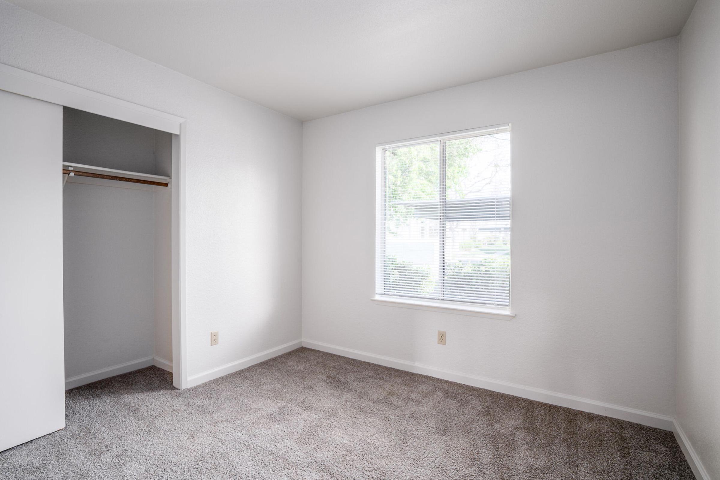 A clean, empty bedroom with light gray carpet and white walls. There is a window on one side letting in natural light and a small closet on the opposite wall. The room appears bright and spacious, suitable for various furnishings or decoration.