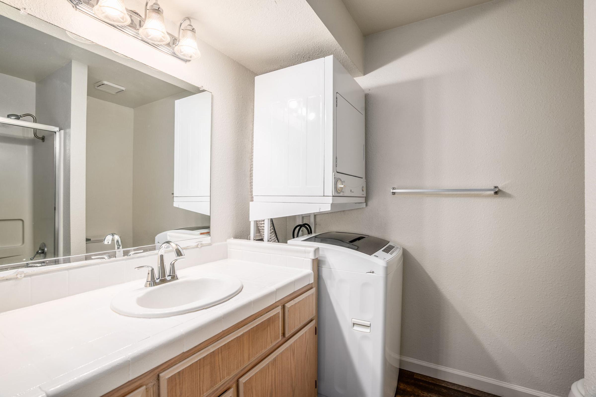 A clean bathroom featuring a white sink with a modern faucet, a large mirror above, a shower area in the background, and a stacked washer and dryer unit. The walls are painted a light color, and the flooring is wood-like. Bright lighting from above enhances the spacious feel.