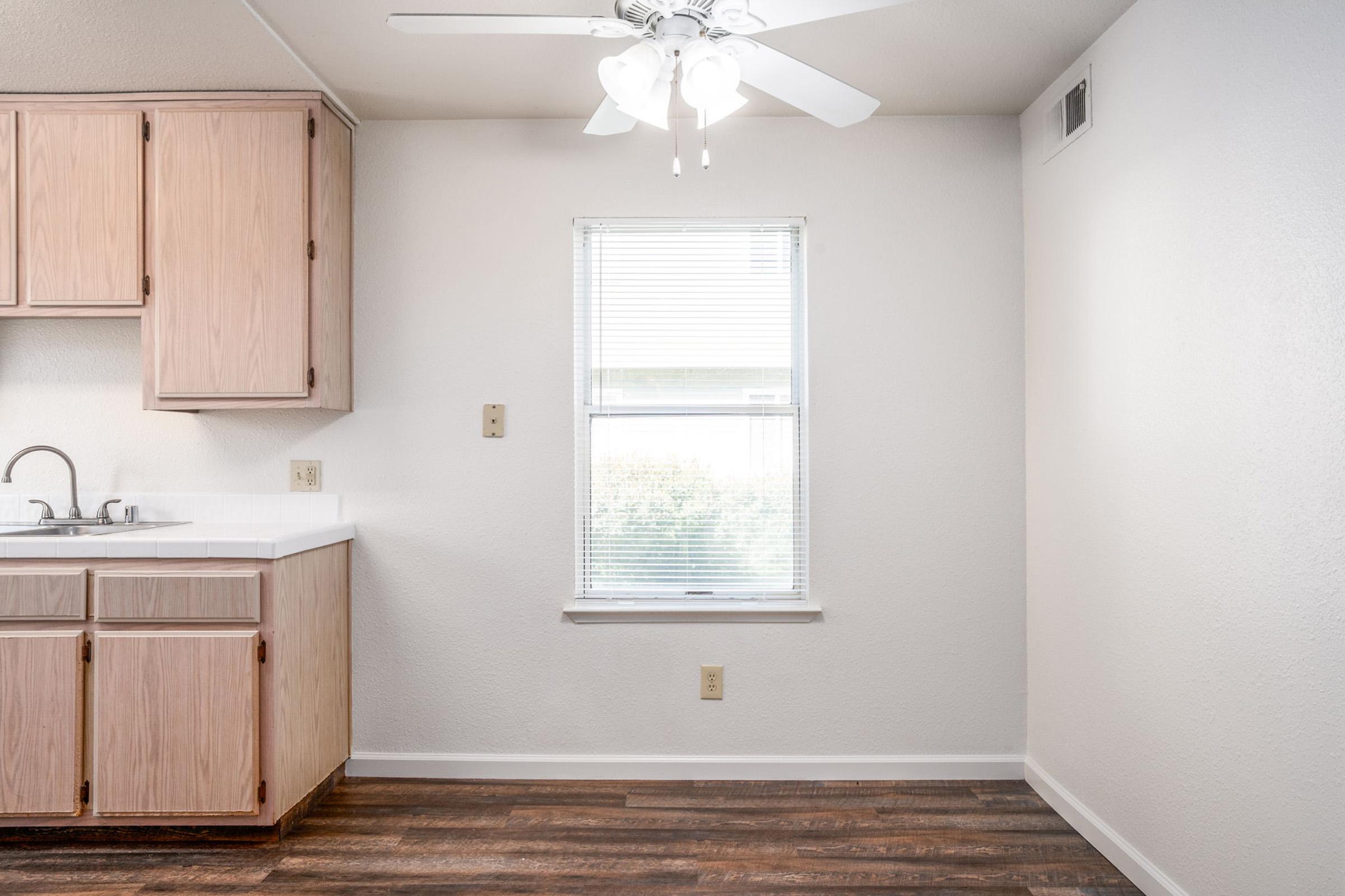 A clean, bright kitchen space featuring light wooden cabinets, a sink with a faucet, and a ceiling fan. Natural light streams in through a window, illuminating the room's simple design and wooden floor. The walls are painted in a neutral color, giving the space a fresh and airy feel.