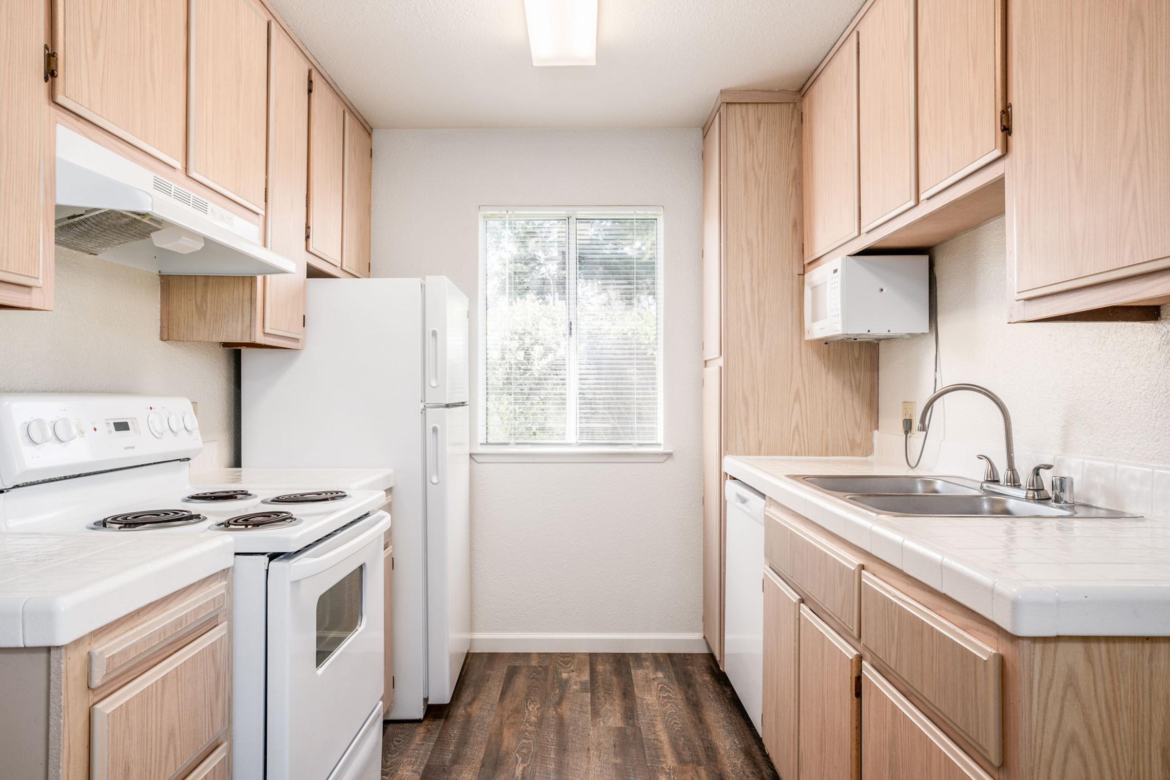 A compact kitchen featuring light wooden cabinets, a white stove and refrigerator, a sink, and a window allowing natural light. The countertops are light-colored, and the flooring is dark wood, creating a contrast with the cabinetry. The kitchen is well-organized and functional.