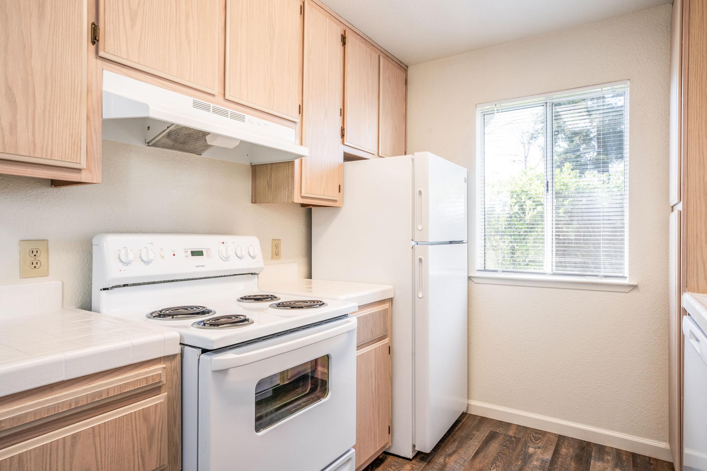 A bright kitchen featuring light wood cabinetry, a white stove with four burners, and a refrigerator. Natural light flows in through a window overlooking greenery. The countertops are white tile, and there's a washer/dryer combo visible in the room. The flooring is dark wood laminate.