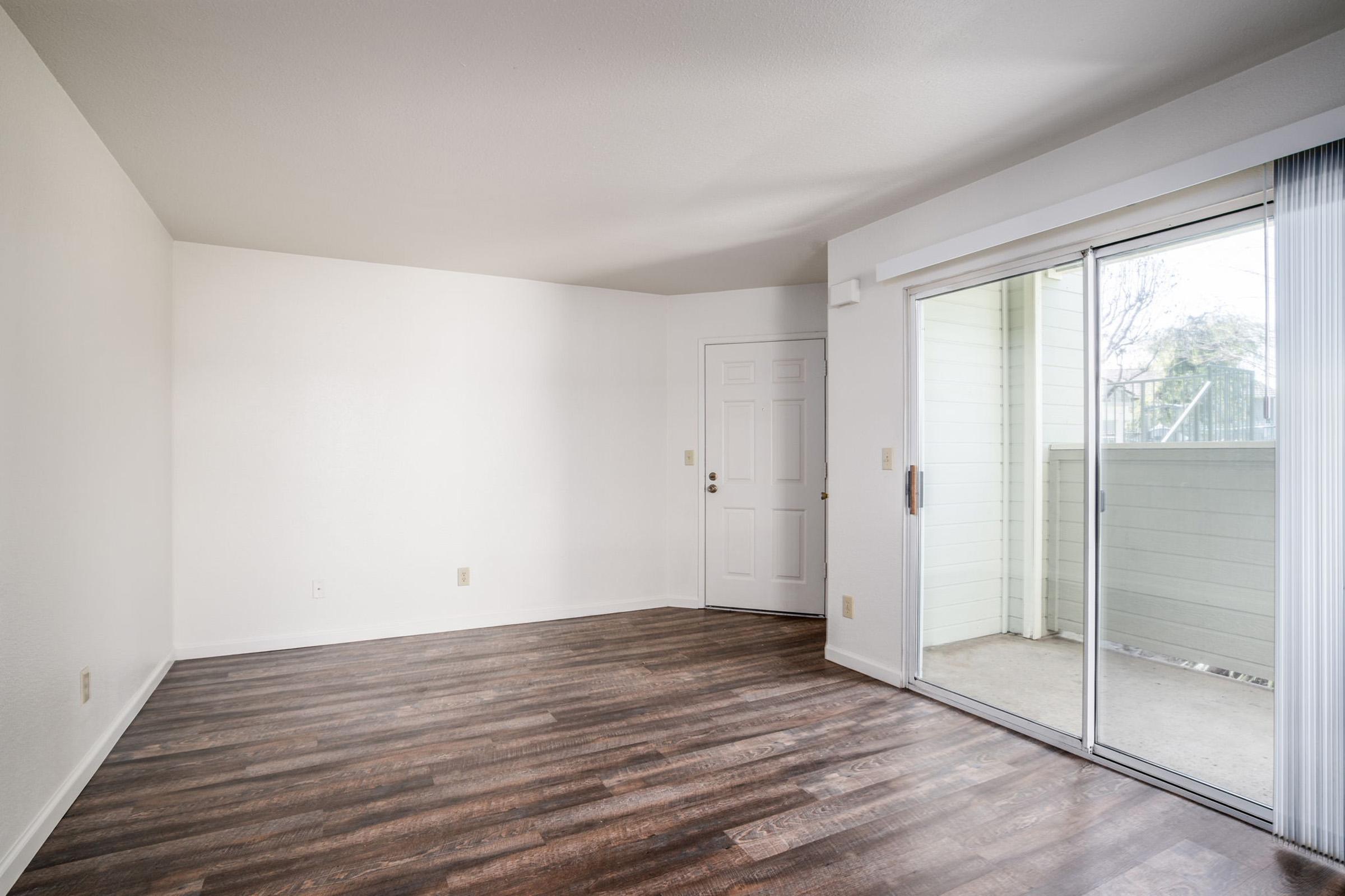 Empty room with light-colored walls and dark hardwood flooring. There is a single door leading to the outside and a sliding glass door with a view of a balcony. Natural light fills the space, creating a bright and open atmosphere.