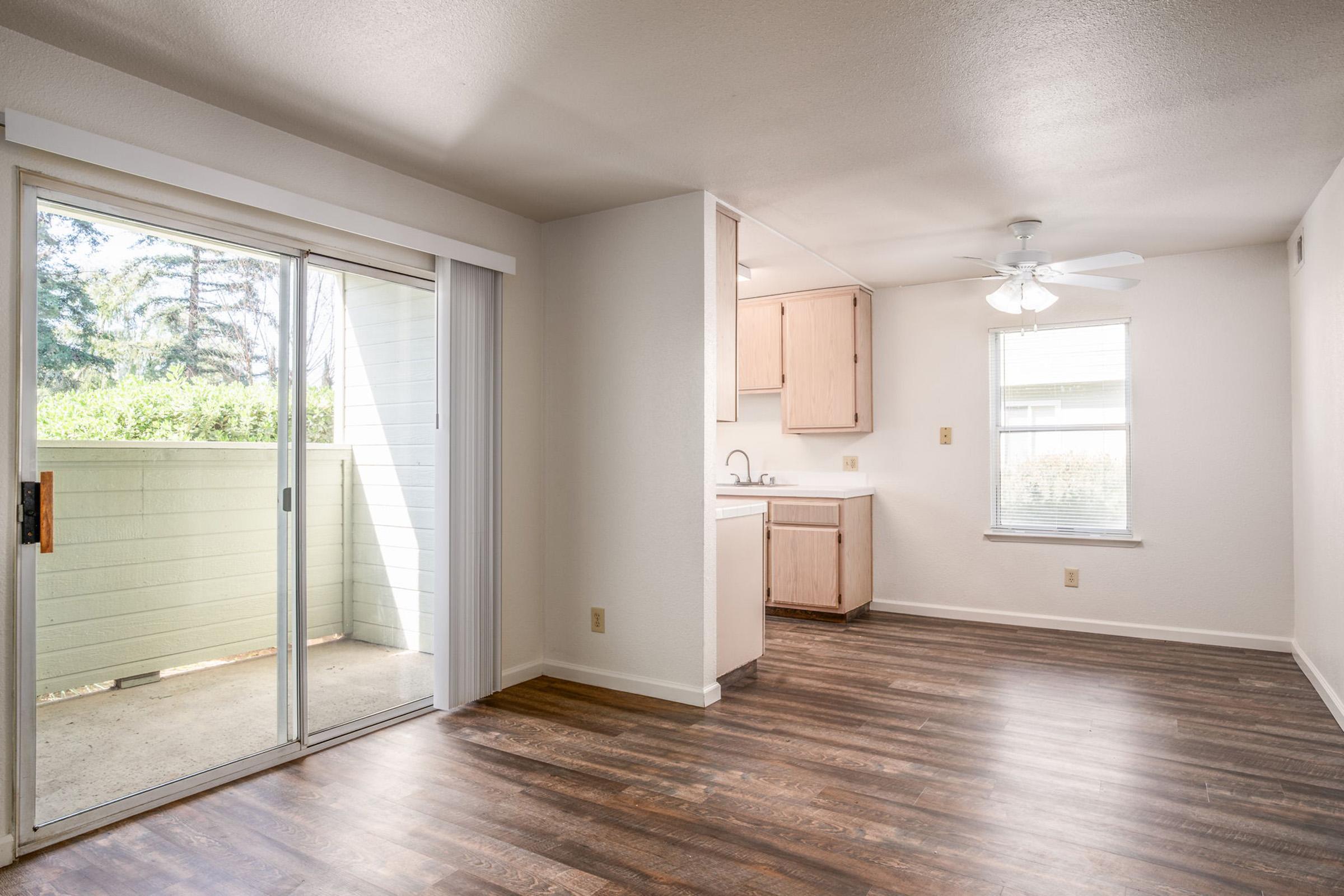 Spacious interior of a light-filled living room with hardwood flooring, sliding glass door leading to a small balcony. A ceiling fan is visible, along with an open kitchen area featuring wood cabinetry. Natural light streams in from the window, enhancing the inviting atmosphere of the room.
