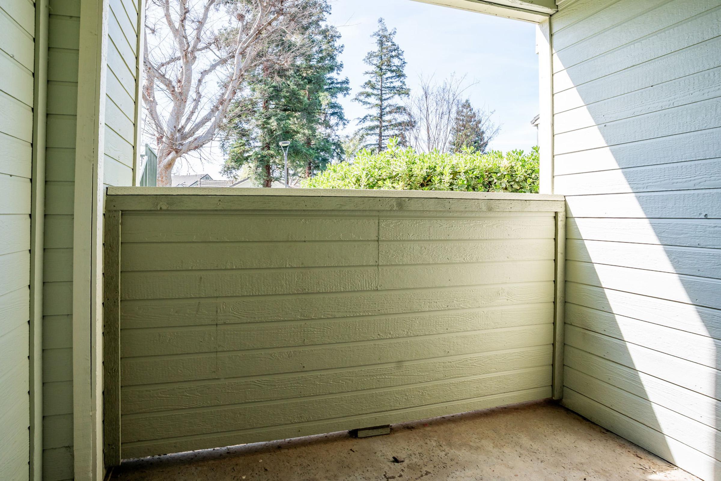 An empty balcony or patio view with light green walls and a wooden railing. Outside, there are trees visible in the background, suggesting a sunny day. The space is well-lit and serene, offering a glimpse of nature beyond the railing.