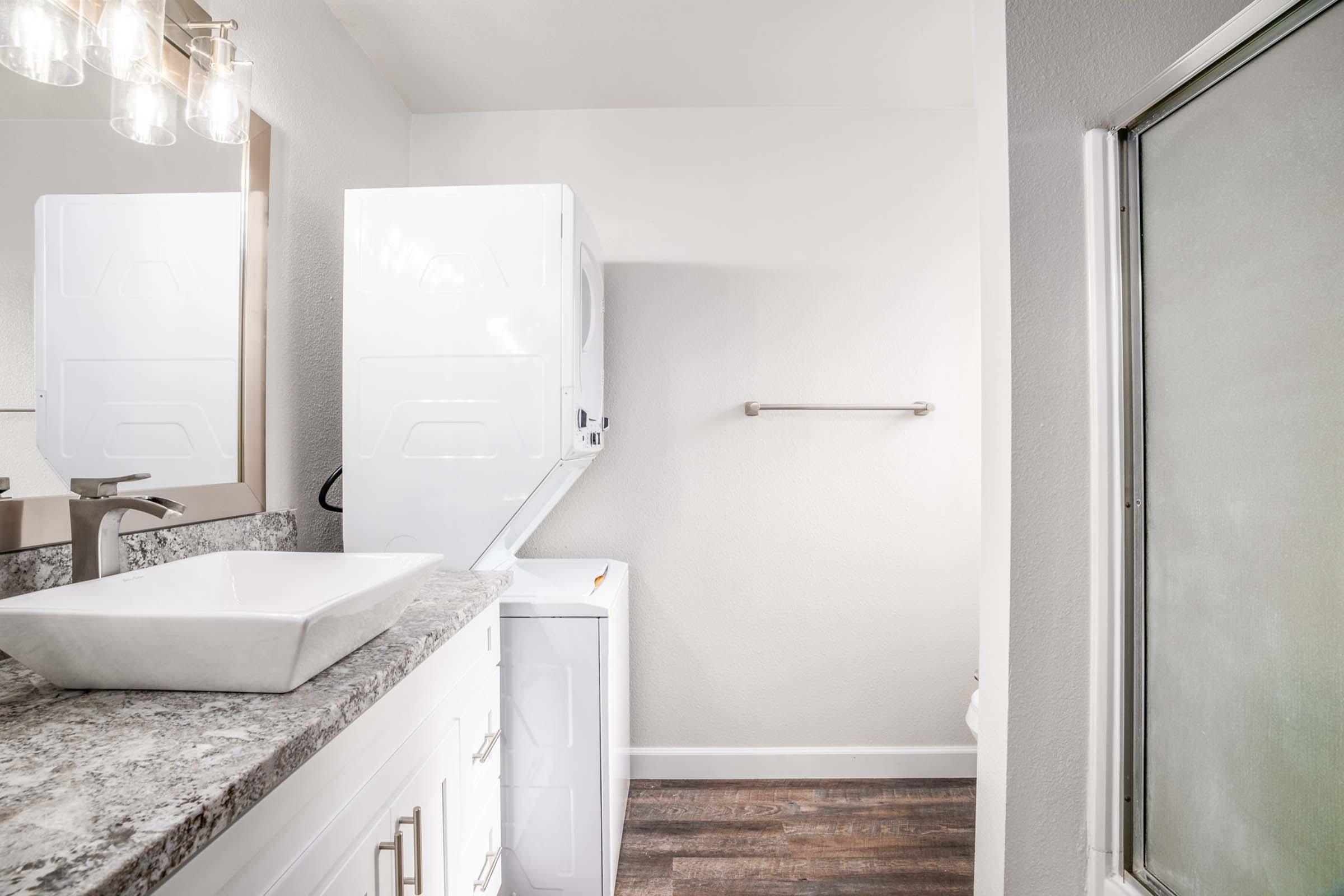 A modern bathroom featuring a countertop with a stylish sink, a washer and dryer stack unit, and a large glass shower. The walls are painted light gray, and there is a towel bar mounted on the wall. The flooring is a dark wood-like material, adding a contemporary feel to the space.