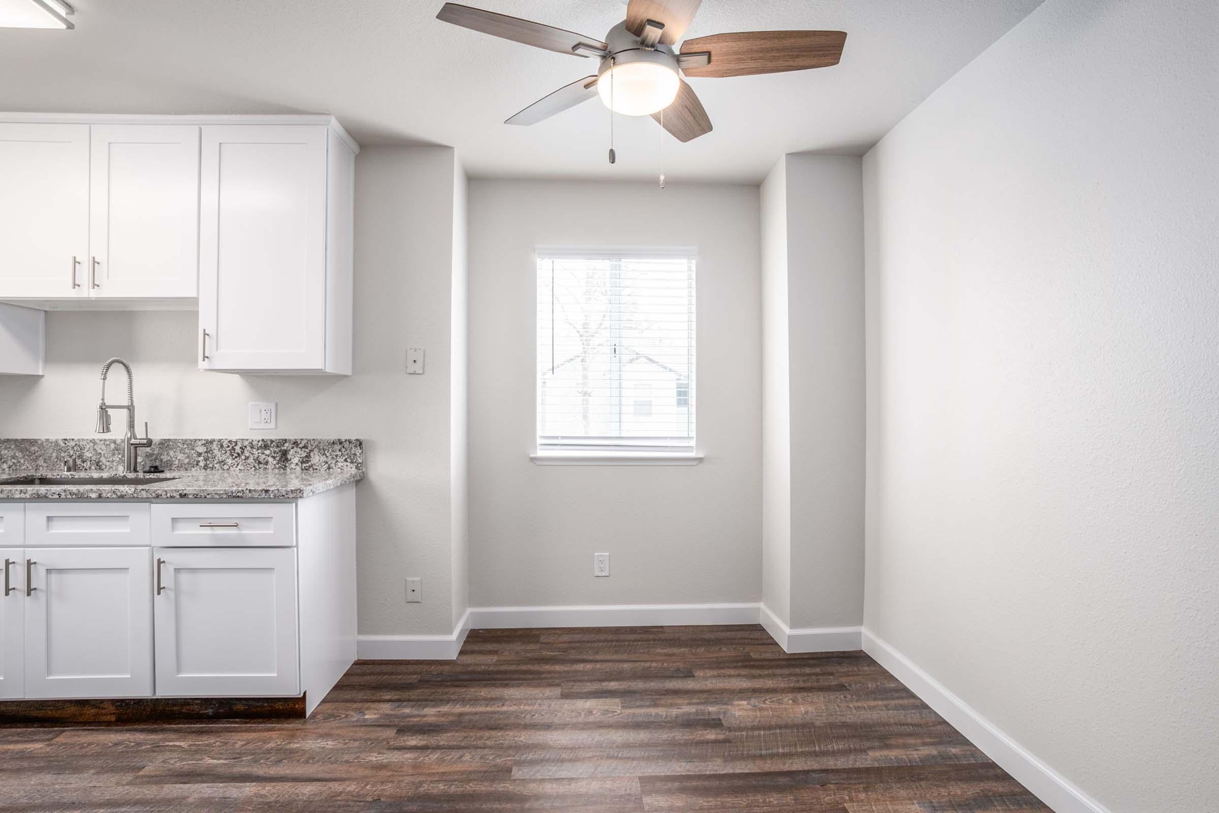 A modern kitchen with white cabinetry, a grey granite countertop, and a stainless steel sink. A ceiling fan with wooden blades hangs above. A window lets in natural light, illuminating the light-colored walls and dark laminate flooring. An empty corner suggests potential for decor or furniture.