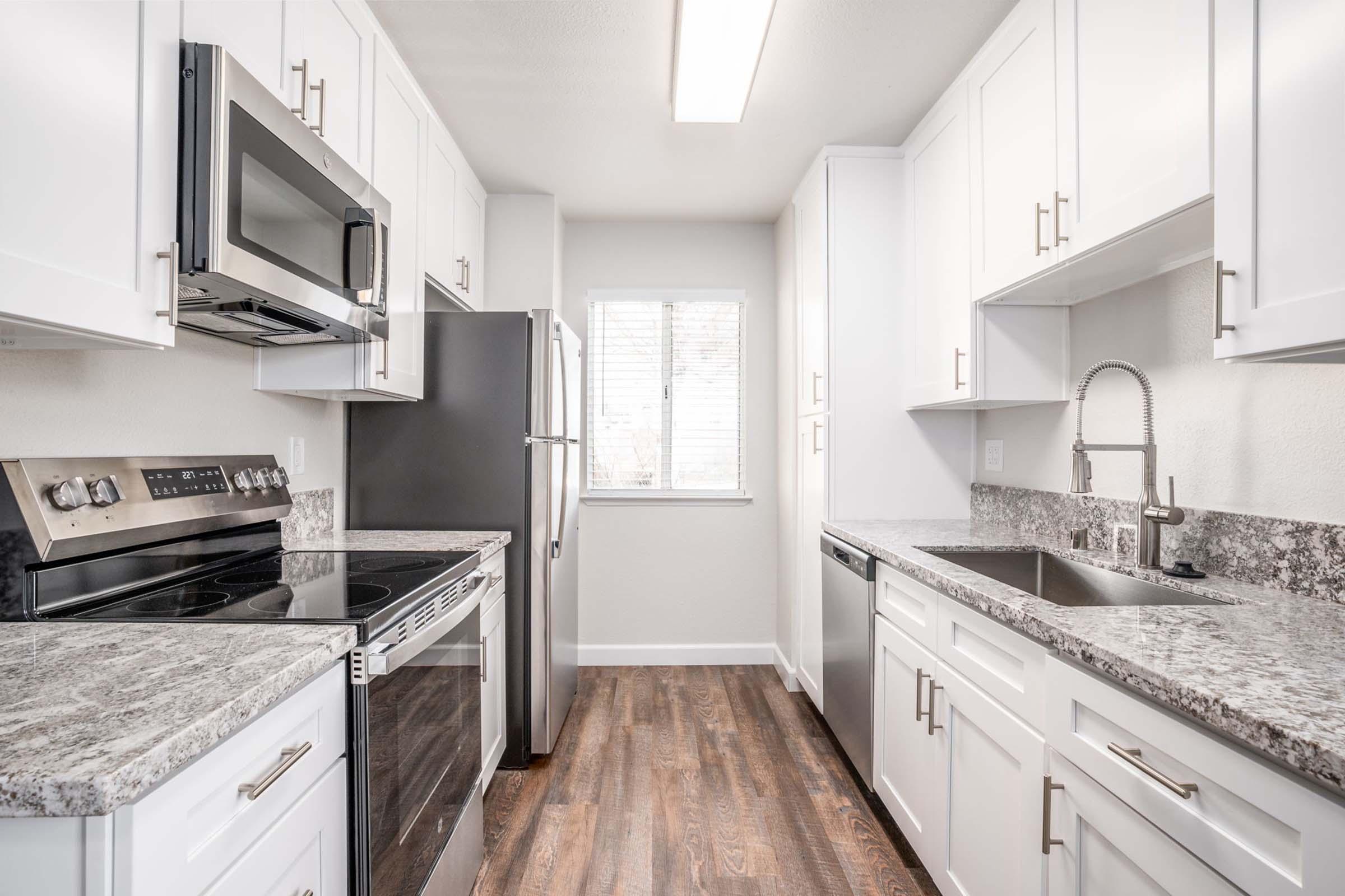 A modern, well-lit kitchen featuring white cabinets, stainless steel appliances, and granite countertops. The layout includes an oven, microwave, refrigerator, and sink, with a window allowing natural light to enhance the space. The flooring is dark wood, adding warmth to the overall design.
