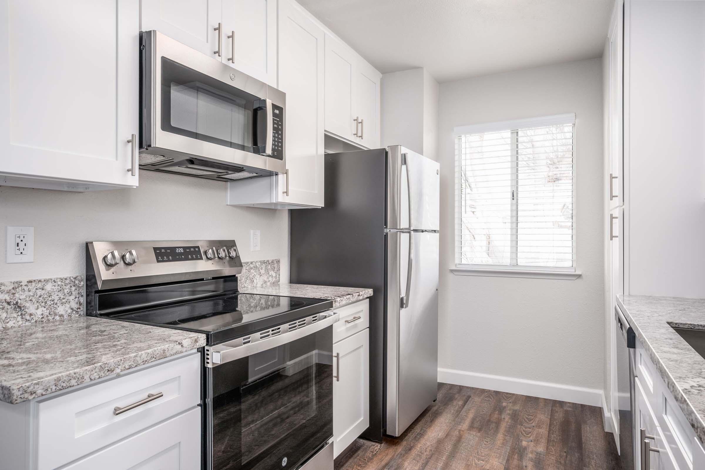 Modern kitchen featuring white cabinets, a stainless steel microwave, an electric stove, and a refrigerator. The countertop has a granite finish, and there is a window allowing natural light into the space, enhancing the bright, clean atmosphere. Hardwood-like flooring completes the contemporary design.