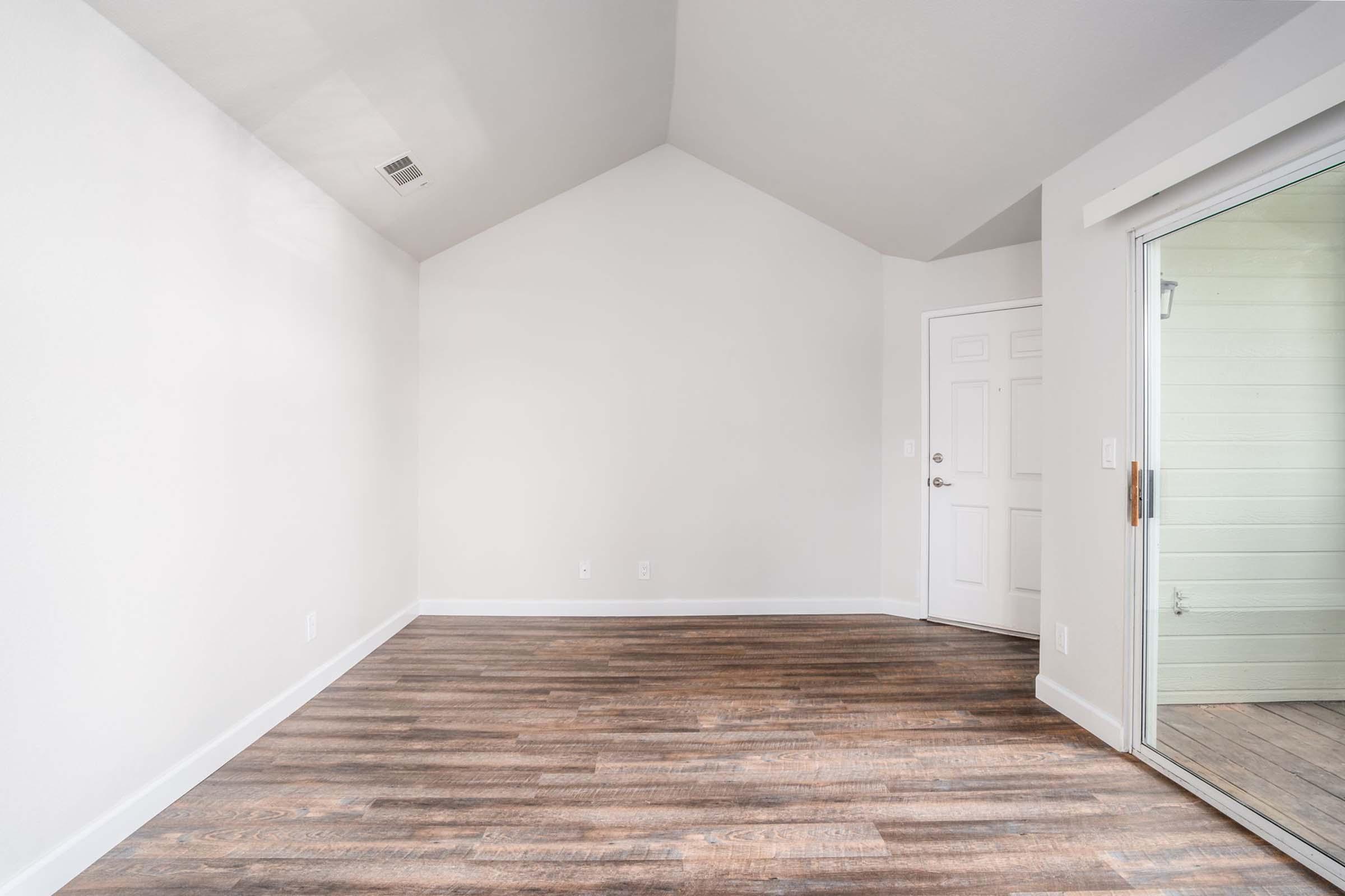 Empty room with wooden flooring, a high ceiling, and plain white walls. A door is visible on the right, leading to a patio or balcony area. Natural light enters through the sliding glass door. The space appears bright and unadorned, ready for decor or furnishings.