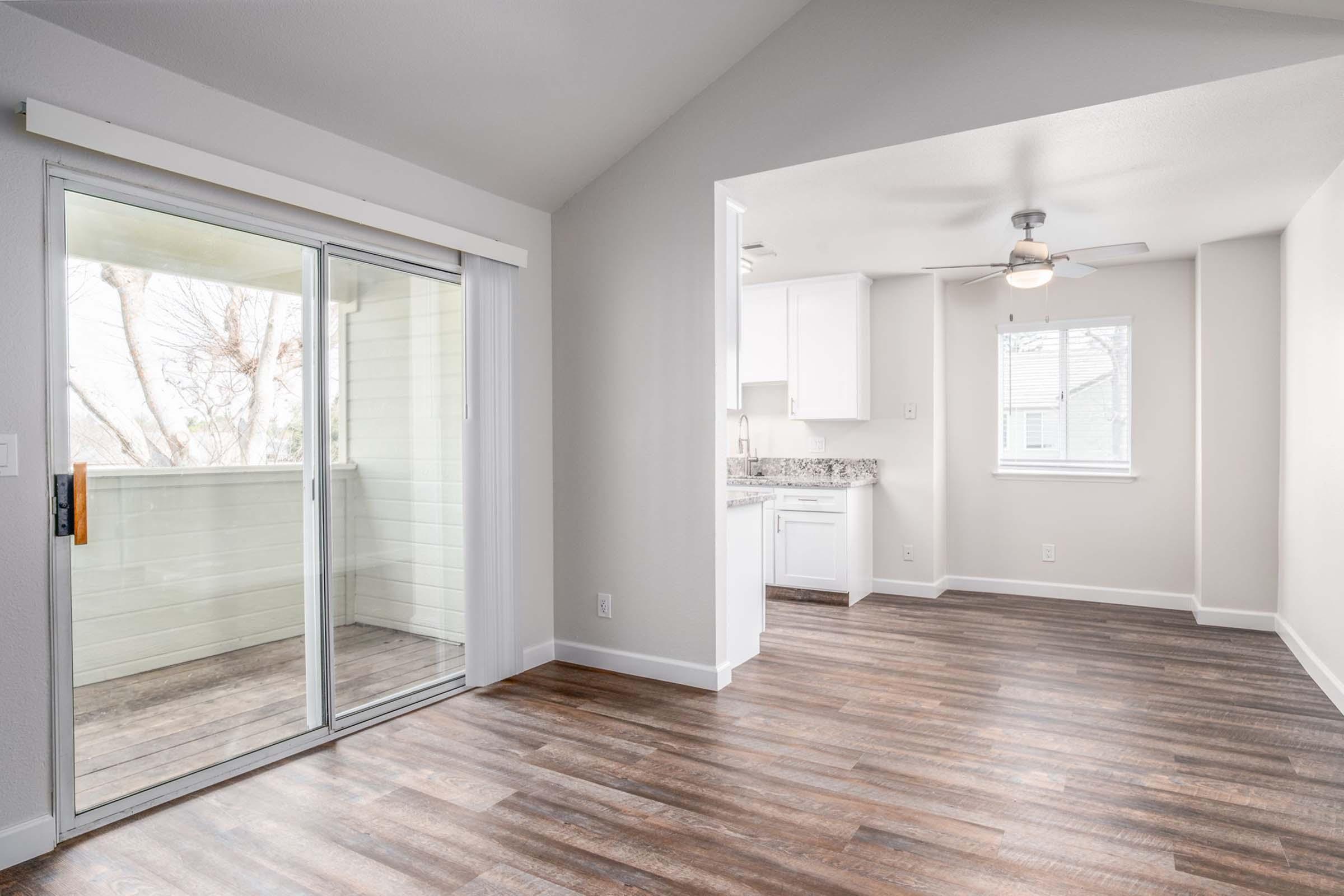 Interior view of a modern living space featuring hardwood floors, white walls, and a ceiling fan. A sliding glass door leads to a small balcony on the left, while a kitchen area is visible in the background, showcasing light cabinets and a window with natural light.