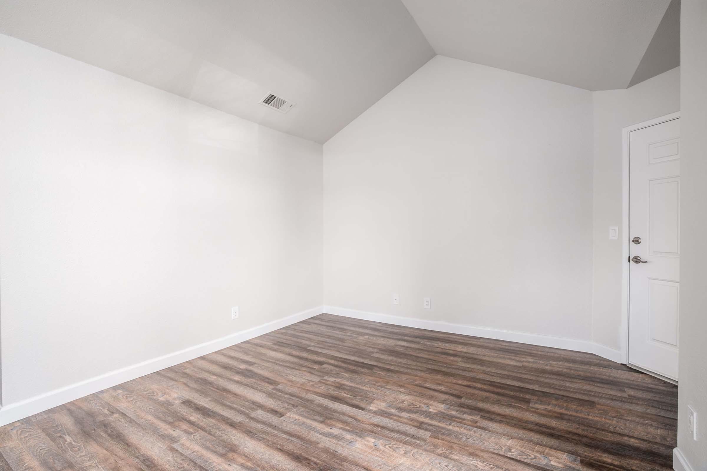 An empty room with a high, sloped ceiling and light-colored walls. The flooring is wood-like laminate, and there is a white door on the right side. The space is well-lit with natural light, creating a bright and airy atmosphere. The room has no furniture or decor, emphasizing its emptiness.