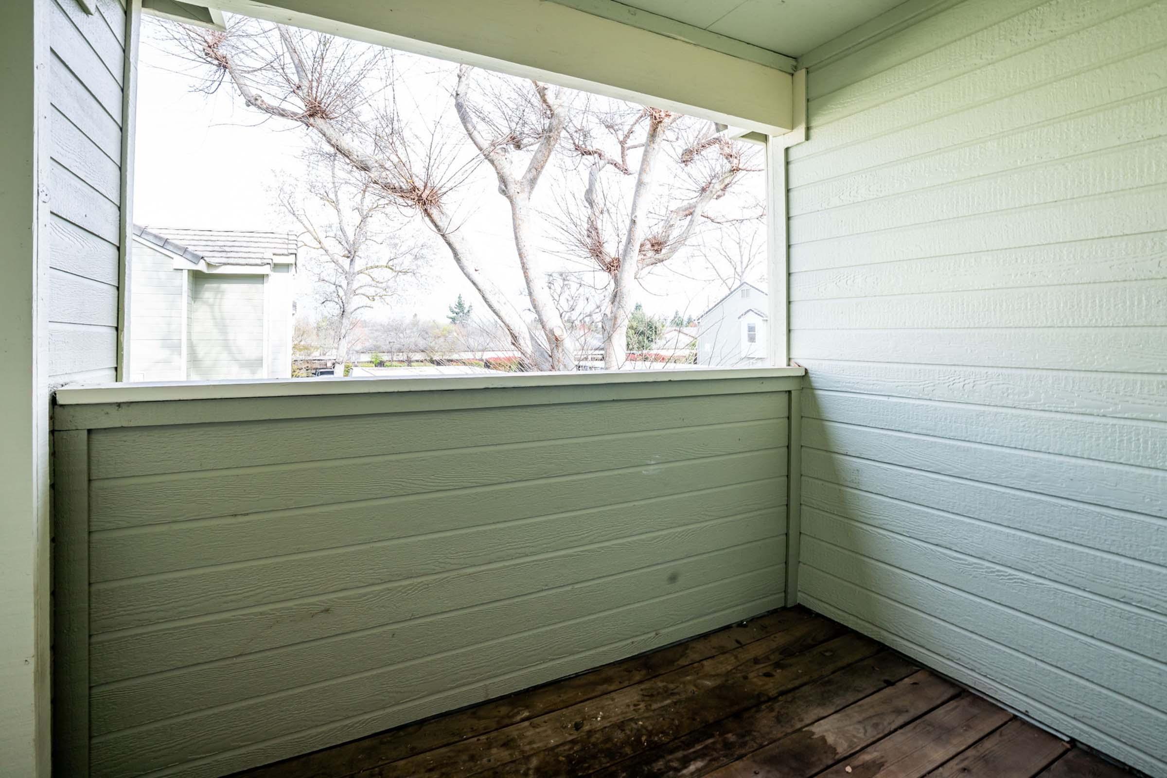 A small, empty porch with wooden flooring and green wooden walls. There's a view of a tree outside the porch, with a soft focus on distant buildings in the background. Natural light fills the space, creating a serene and open atmosphere.