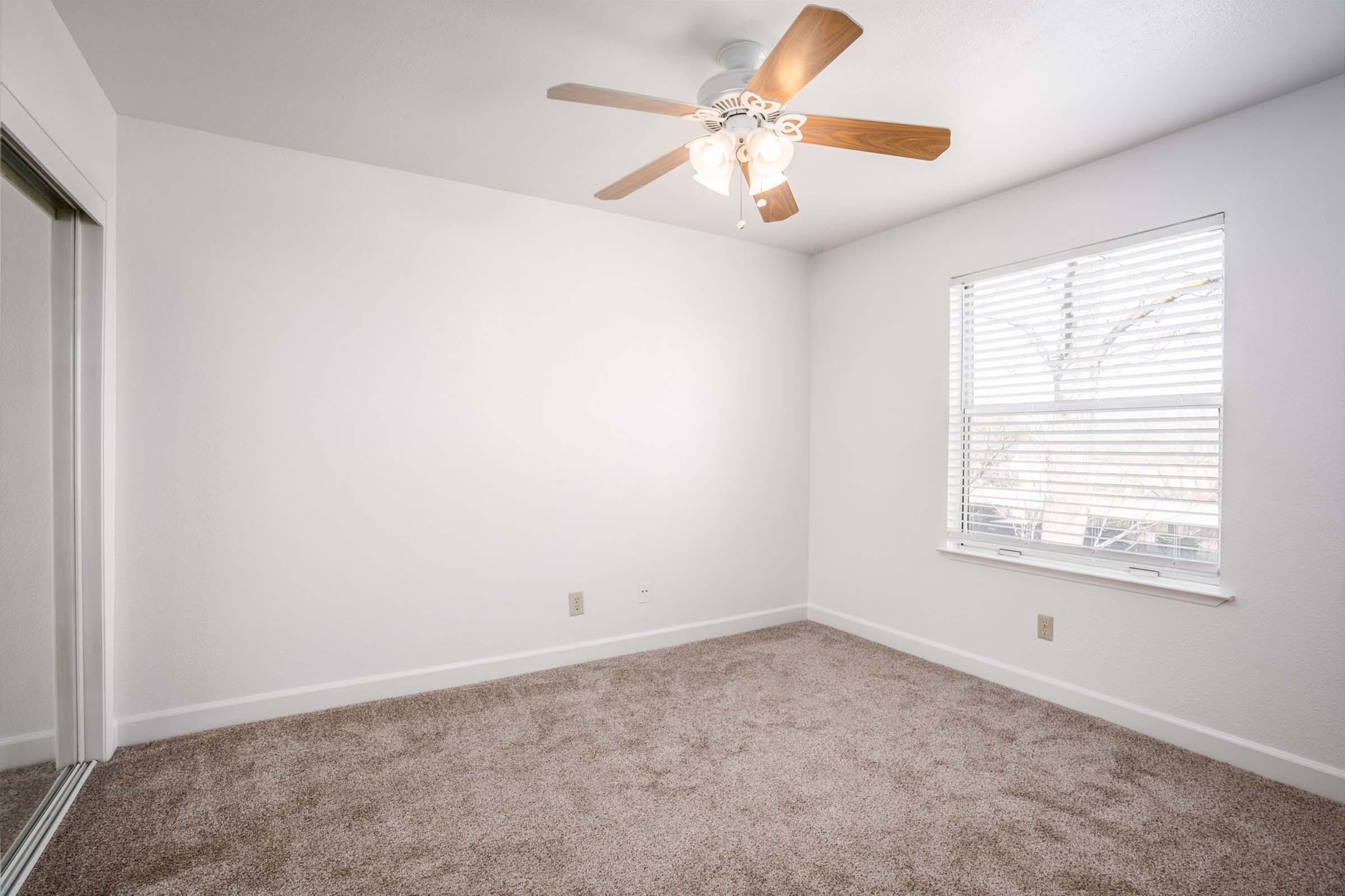 A clean, empty bedroom featuring light beige carpet, white walls, a ceiling fan with wooden blades, and a window with blinds letting in natural light. The room has a simple and airy feel, making it suitable for various interior design options.
