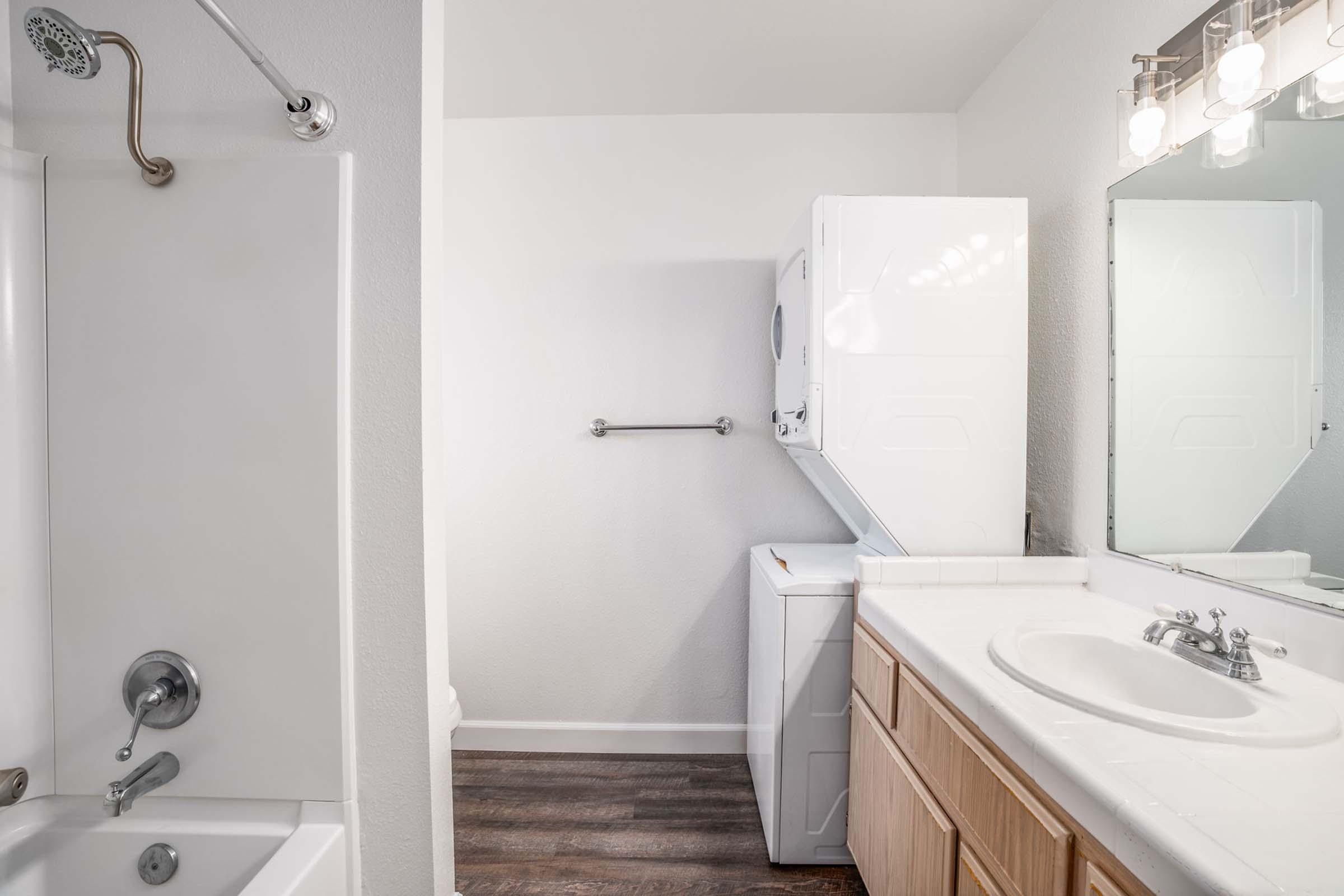 A clean, modern bathroom featuring a white bathtub and shower, a large mirror above a sink with wooden cabinetry, and a stacked washer and dryer unit. The walls are painted light, and the flooring is a dark, wood-like laminate.
