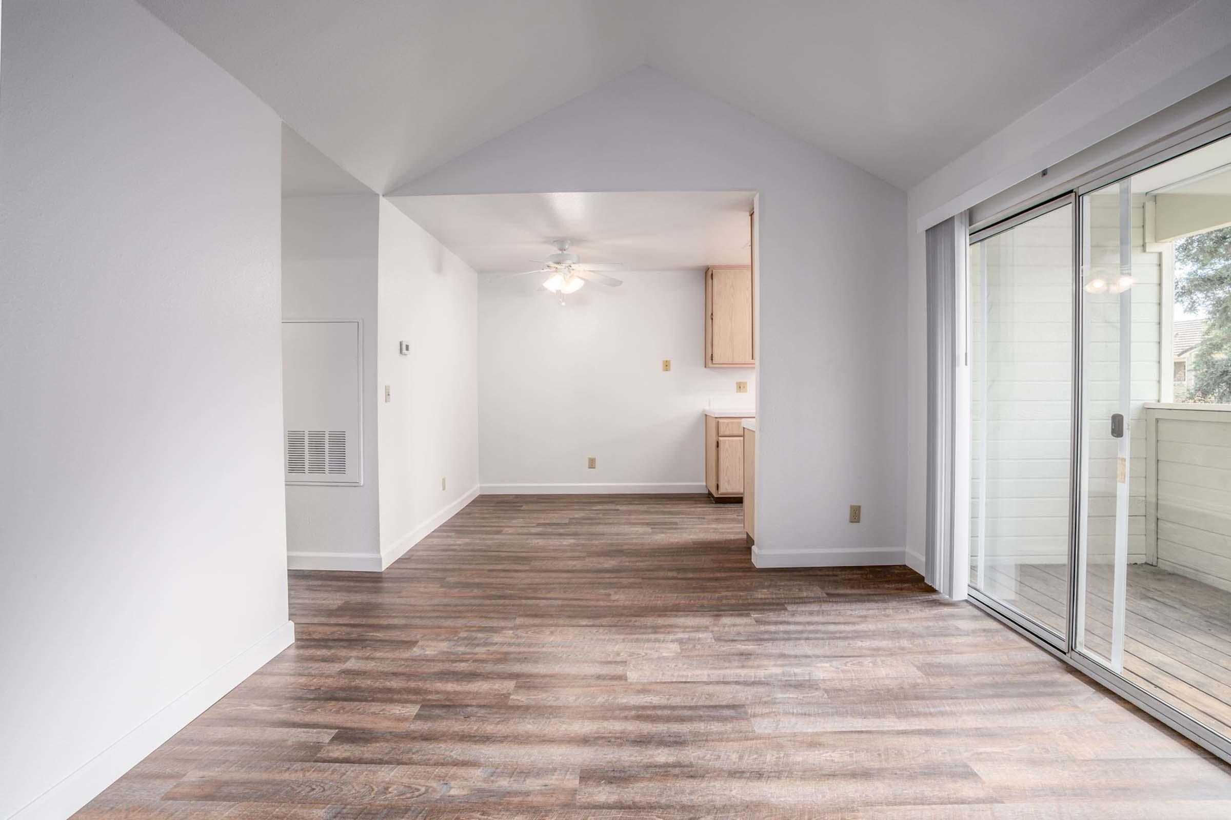 An interior view of a bright, open-concept living space with light wood-like flooring, a white ceiling, and a ceiling fan. It features a large sliding glass door leading to a balcony, and an adjacent kitchen area visible in the background with wooden cabinets. The walls are painted in a neutral color.