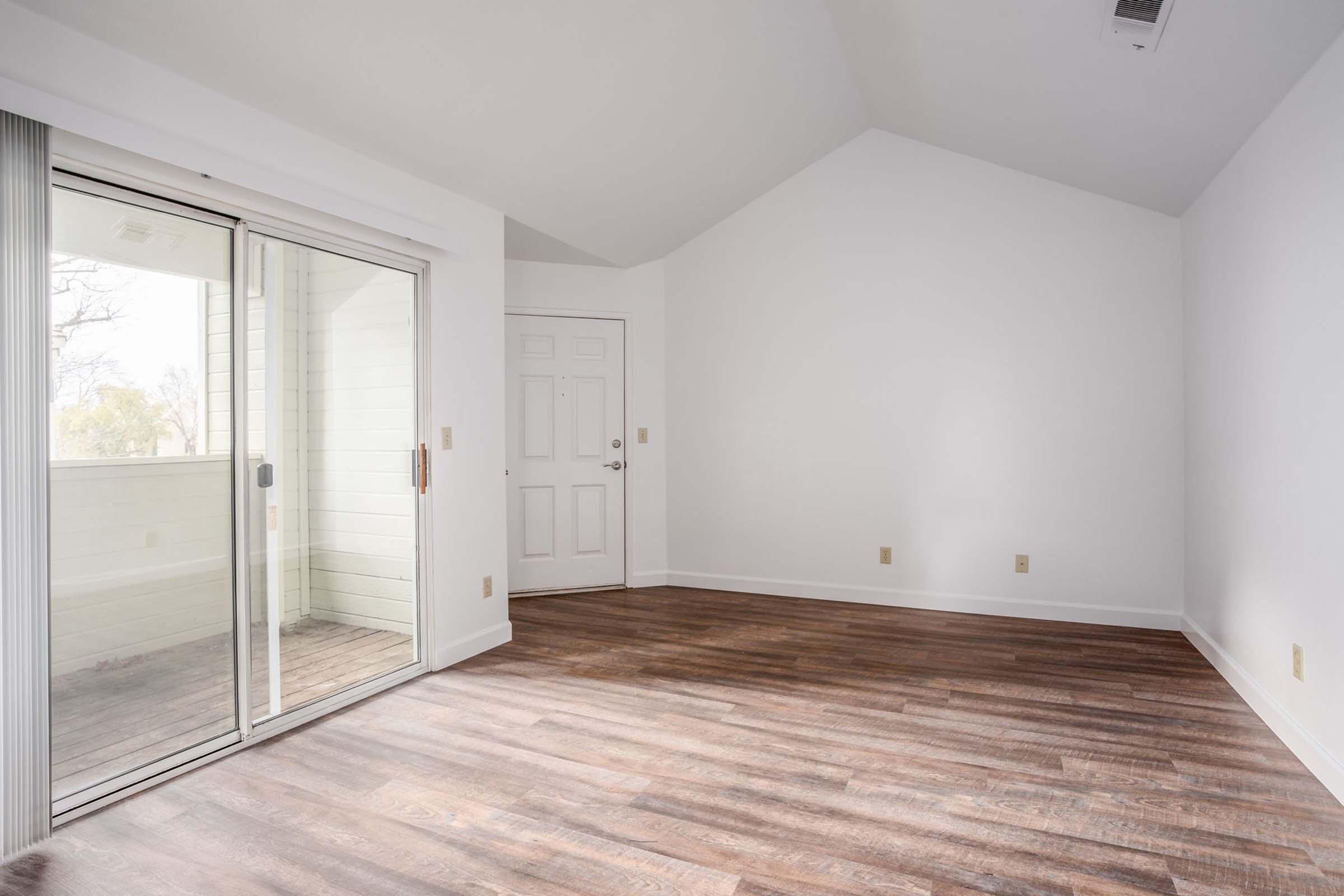A bright, empty room with a high ceiling, featuring a sliding glass door that opens to a balcony and a standard door on the right. The floor is made of dark wood, and the walls are painted white, creating a spacious and airy atmosphere. Natural light floods the space from the balcony.
