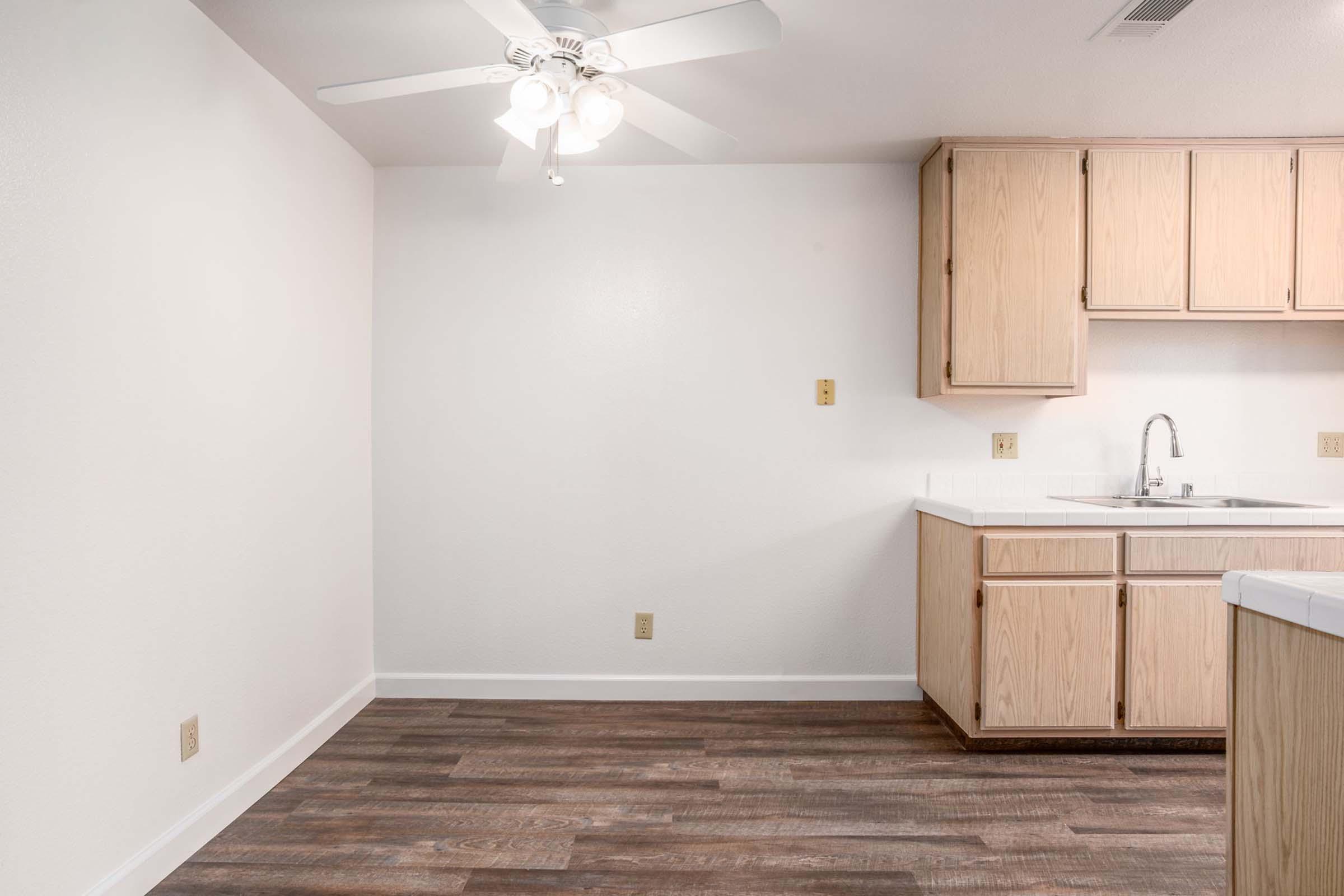 A clean, empty kitchen featuring wooden cabinets, a sink, and a ceiling fan. The walls are white, and the floor has dark wooden planks. The space is well-lit and has a minimalist design.