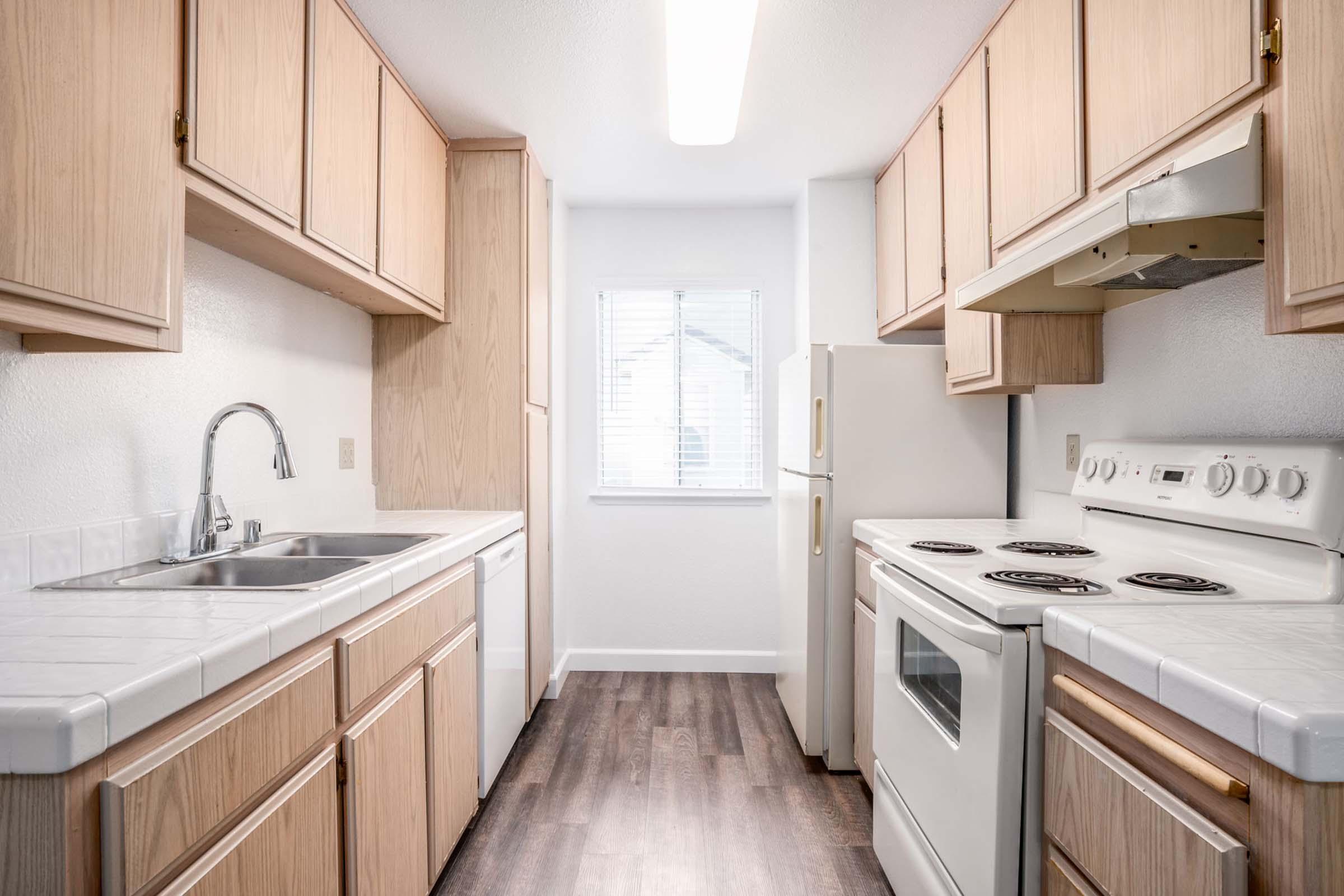 A modern kitchen featuring light wood cabinetry, a double sink, a white refrigerator, and a stove with an oven. The space has a window allowing natural light in, and the flooring is a dark wood laminate, creating a contrast with the light-colored cabinets and countertops.
