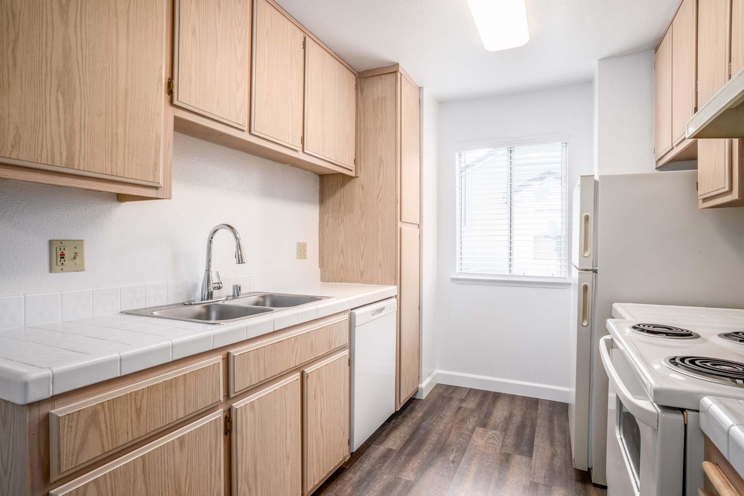 Light and bright kitchen featuring wooden cabinets, a sink with a modern faucet, a dishwasher, and an electric stove. The countertops are white and tiled, with a window providing natural light. The floor is dark-colored laminate, creating a warm contrast with the cabinetry.