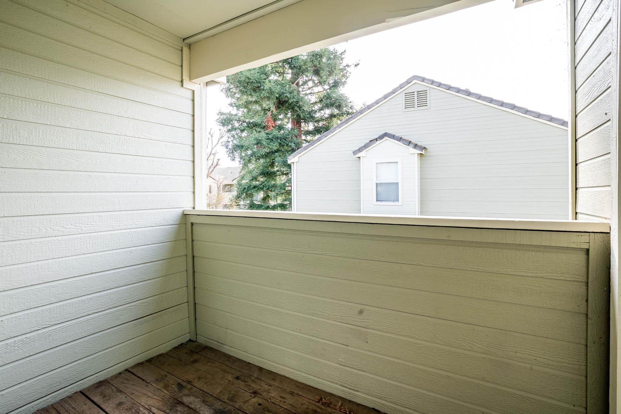 A view from a porch or balcony showcasing a wooden floor and light-colored wooden walls. In the background, a green tree and a light gray house with a peaked roof are visible. Natural light illuminates the scene, creating a serene outdoor space.