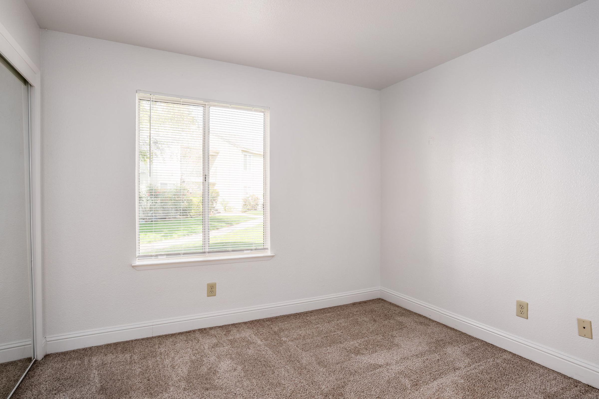 A small, empty room with light beige carpet and white walls. It has a window with blinds that lets in natural light, and a mirrored closet on one wall. The view outside shows greenery. The room is unfurnished and has a simple, clean appearance.