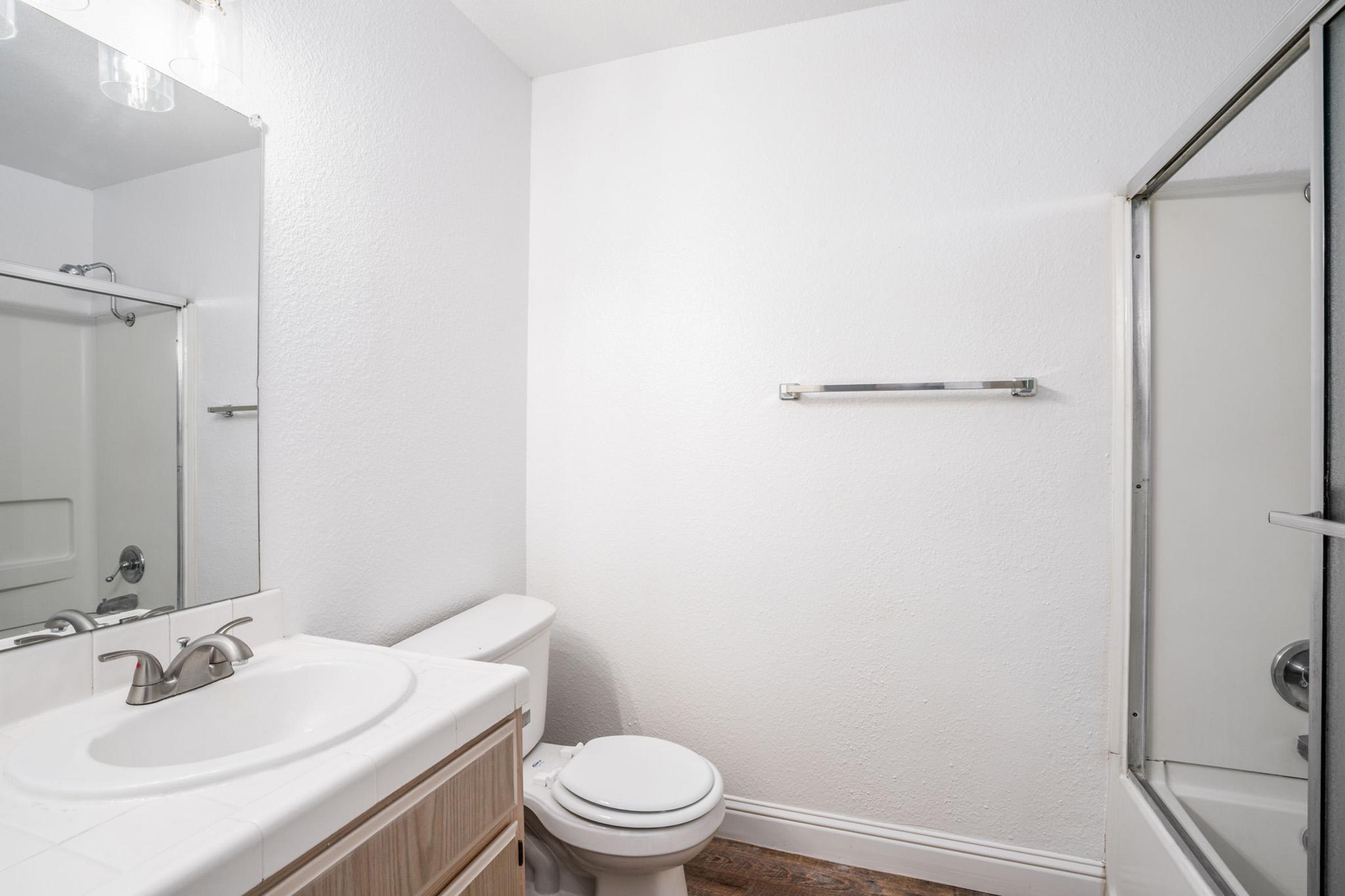 A clean, modern bathroom featuring a white wall, a large mirror above a sink with a wooden cabinet, a toilet, and a glass shower stall. There is a towel rack on the wall and wooden flooring, creating a bright and minimalistic atmosphere.