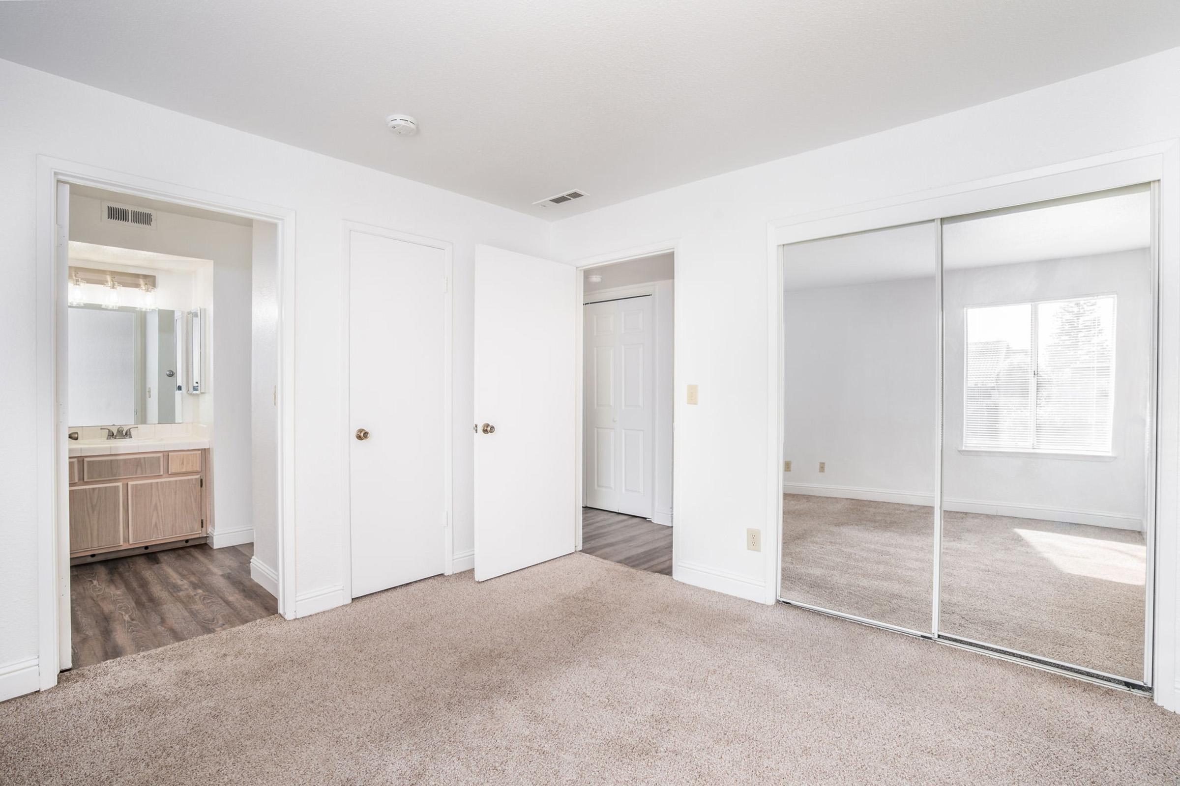 A spacious, well-lit bedroom featuring neutral-colored walls and plush carpet. The room includes a large mirrored closet, two doorways leading to other areas, and a window allowing natural light to brighten the space. A bathroom is visible through a doorway, adding convenience to the layout.