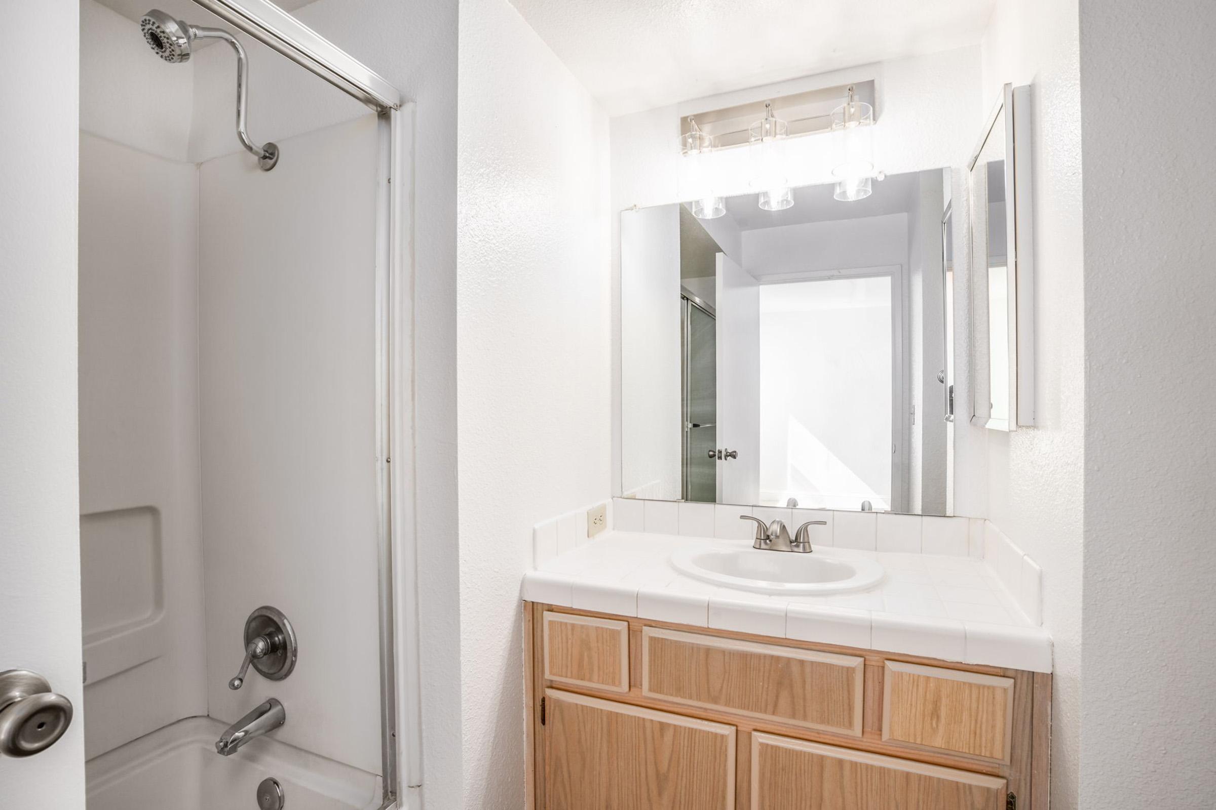 A bright, modern bathroom featuring a shower in one corner, a vanity with a sink, and a large mirror above the sink. The walls are painted in a clean white color, creating a spacious and airy feel. Natural light enters through a window, illuminating the space. The wooden cabinetry adds warmth to the design.