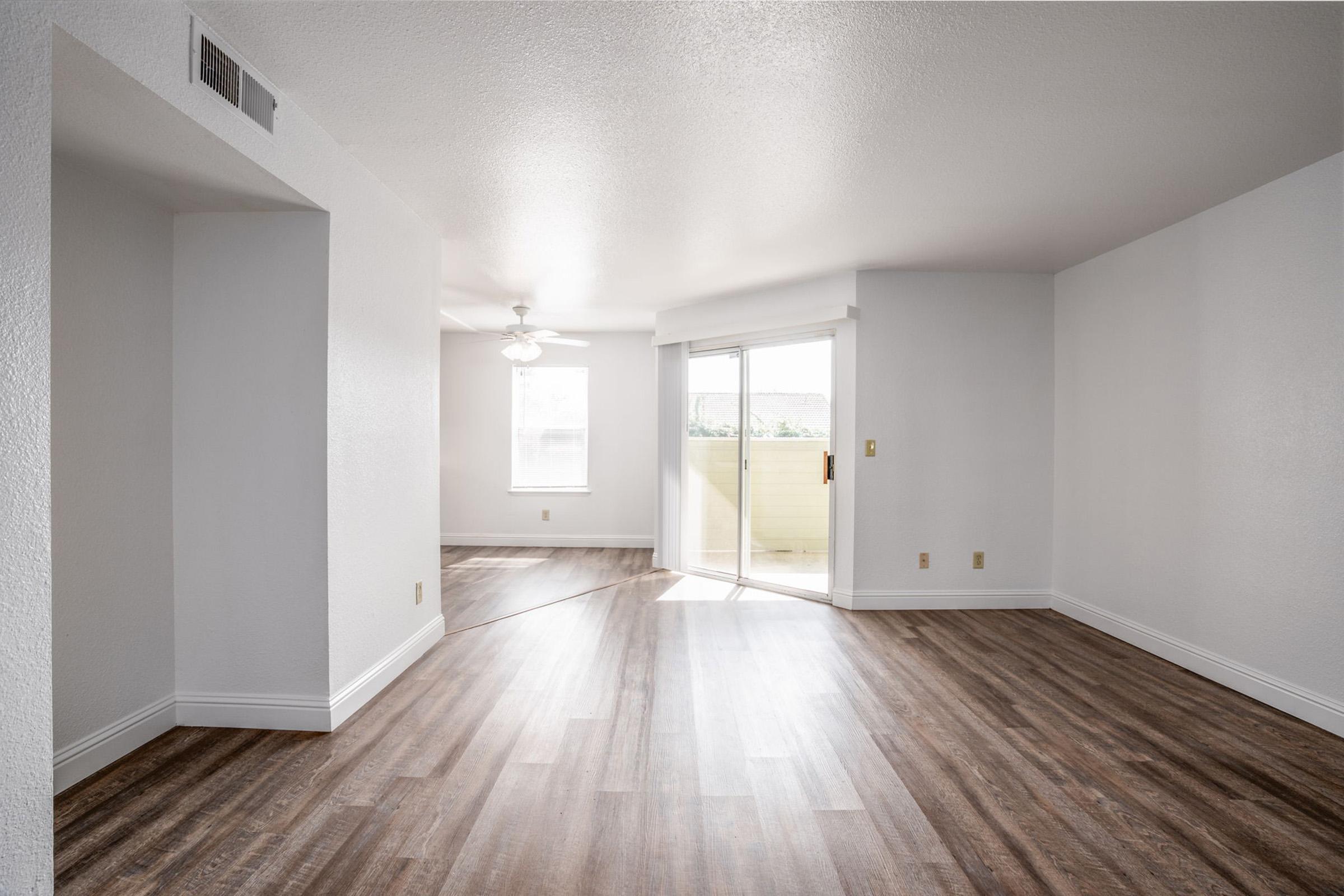 A spacious, empty room featuring light-colored walls and polished wood-like flooring. Natural light floods in from a large sliding door that leads to a balcony, while a ceiling fan hangs in the corner. The room is clean and modern, providing a blank canvas for decoration and furnishing.