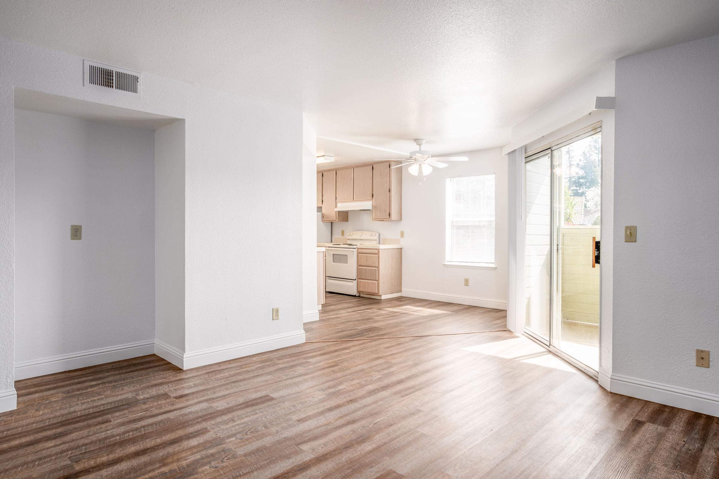 Spacious, bright living area featuring light-colored walls, hardwood flooring, and a ceiling fan. An adjoining kitchen with light wood cabinetry is visible, and there is a sliding glass door leading to a patio or balcony, allowing natural light to fill the space.