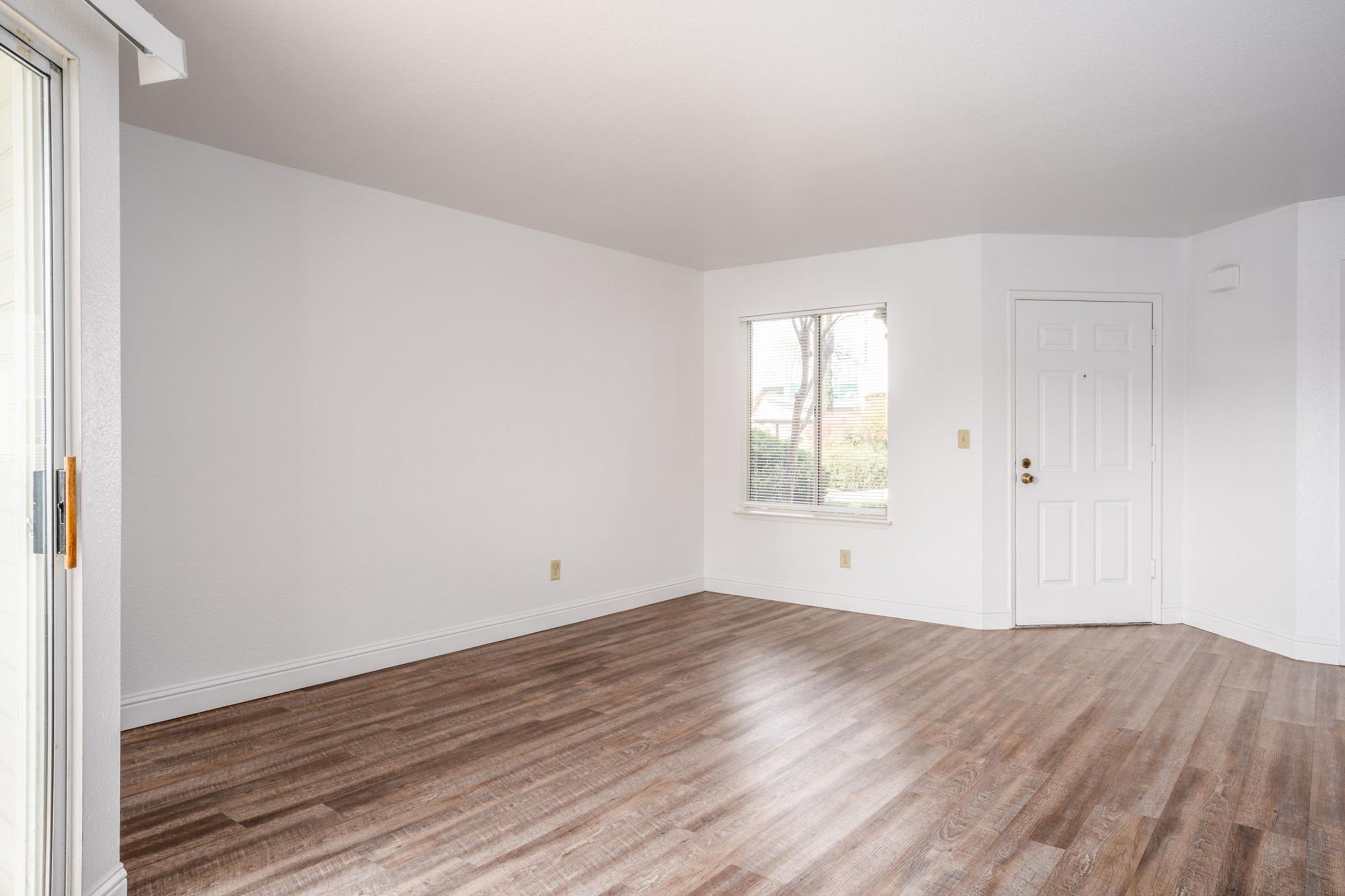 Empty room with light-colored walls and a large window allowing natural light. The floor is covered with wood-like laminate flooring. There is a door on the right side leading outside. The space is clean and unfurnished, creating an open and inviting atmosphere.