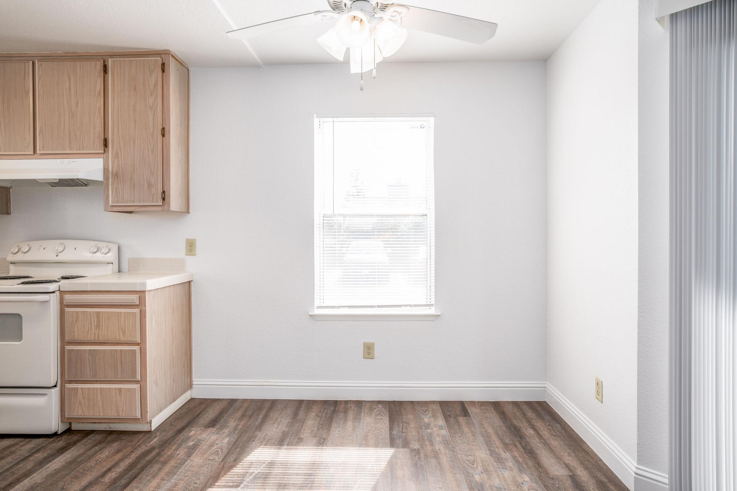 Bright, airy kitchen area featuring light wood cabinetry, a white stove, and a ceiling fan. A large window allows natural light to flood the space, highlighting the wood-look flooring. The walls are painted white, creating a clean and modern atmosphere.