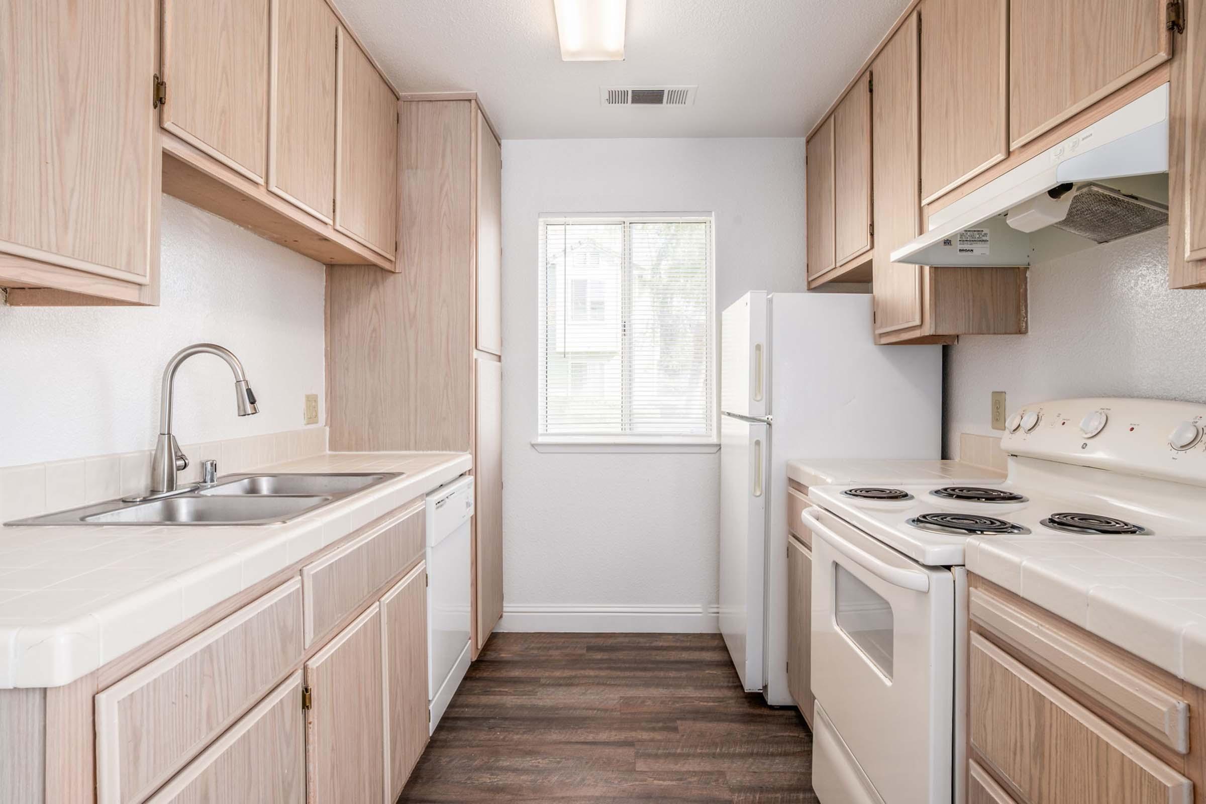 Bright, modern kitchen featuring light wood cabinets, a double sink, a dishwasher, and a white stove. A window provides natural light, and the floor is dark laminate. The kitchen is clean and uncluttered, with ample storage space and a functional layout.