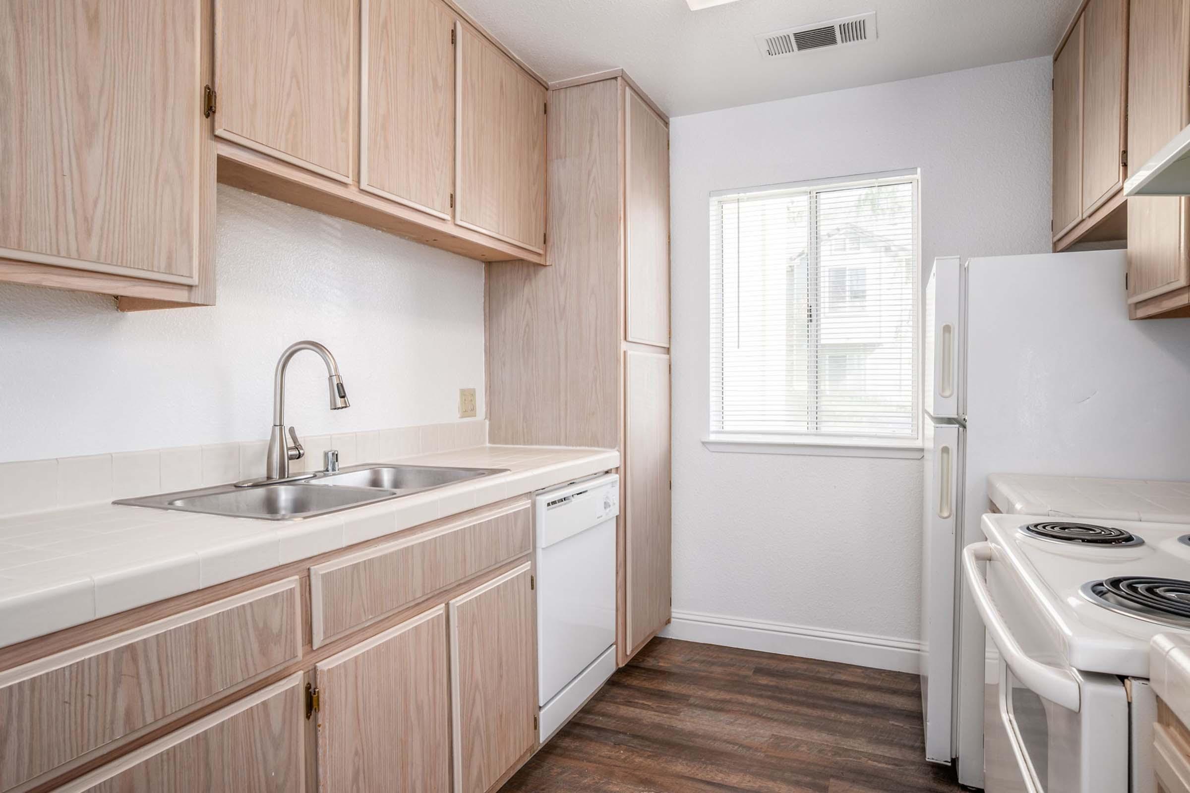 Bright kitchen with wooden cabinets, white countertops, and appliances including a dishwasher and stove. There is a window providing natural light, and the flooring is dark wood. The overall design is modern and functional, suitable for cooking and meal preparation.