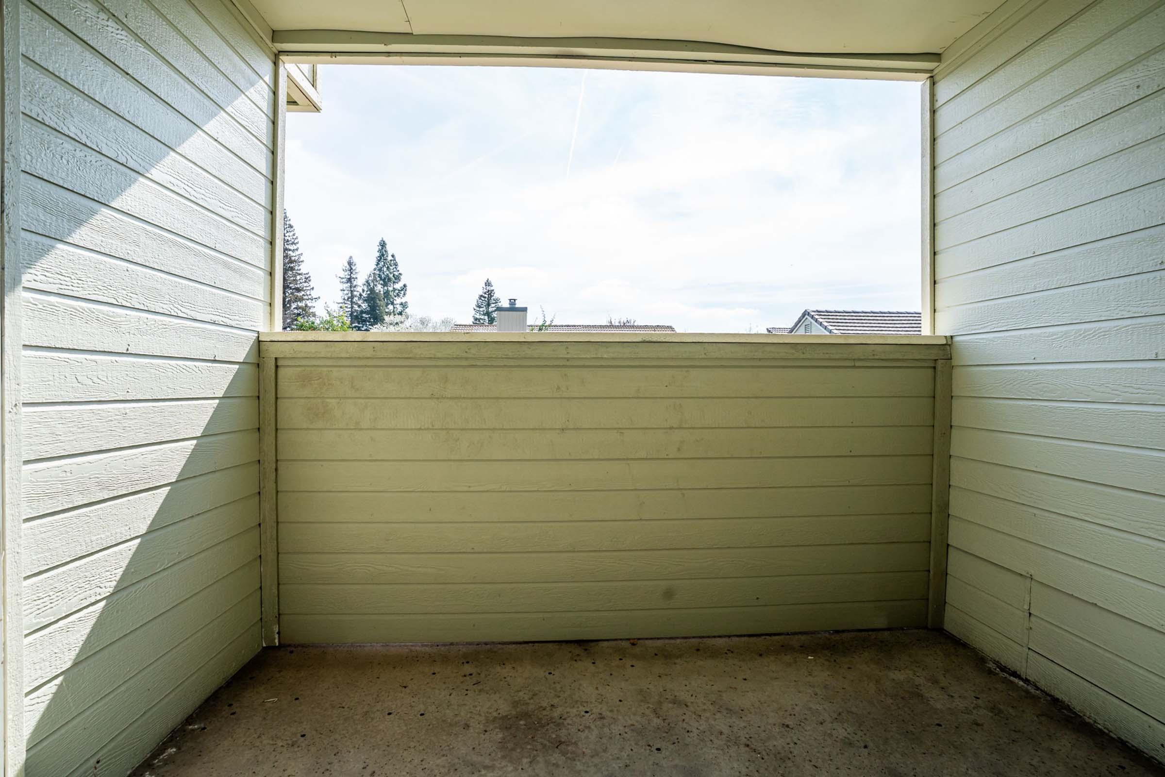 An empty balcony or porch with light green wooden walls and a concrete floor, featuring a wide opening that provides a view of the outside. In the background, there are trees and a clear sky, creating a sense of openness and natural light.