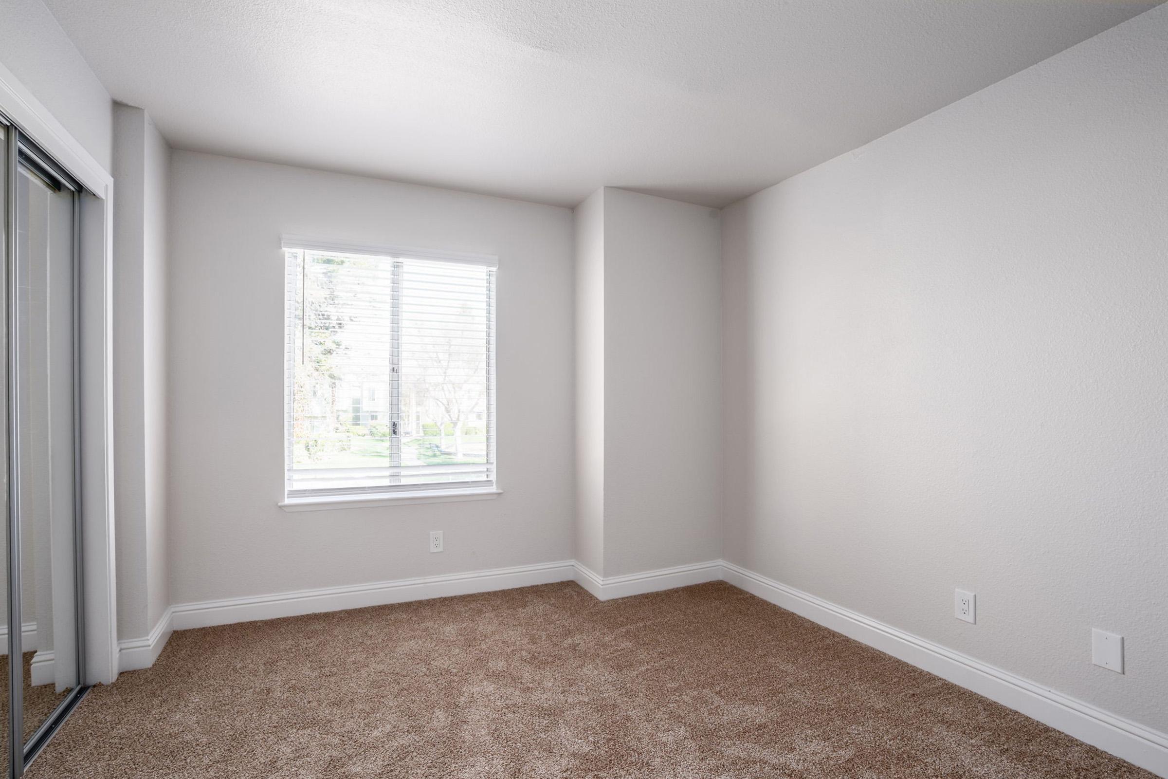 A light, airy room with beige carpet and neutral-colored walls. It features a window with blinds that lets in natural light, a corner alcove, and a mirrored closet door, creating an open and spacious feel.