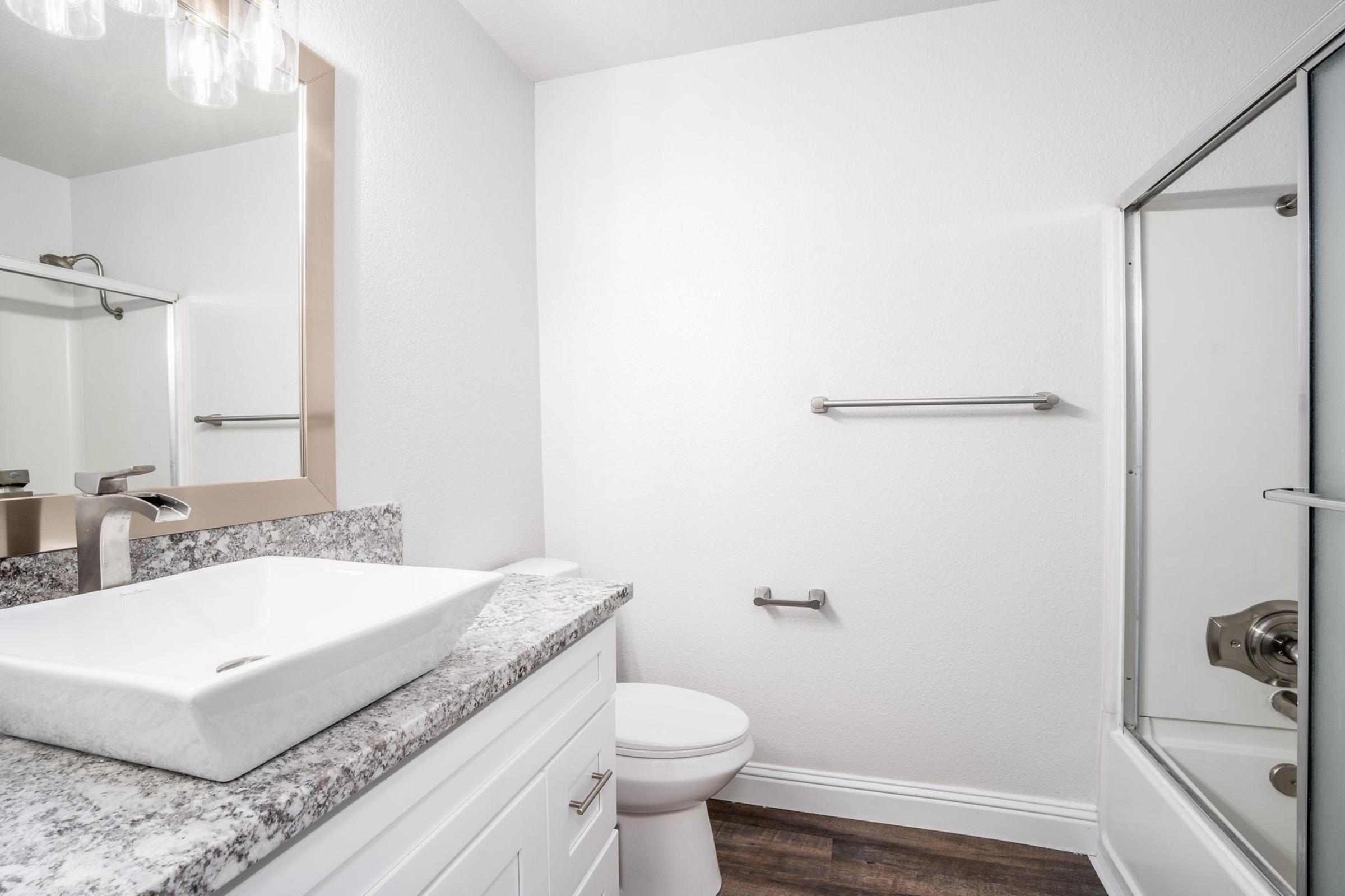 Modern bathroom featuring a white sink with a granite countertop, a wall-mounted mirror, and a toilet. The room has a glass shower enclosure and a towel rack. The walls are painted white, and the flooring is dark laminate, creating a clean and contemporary aesthetic.