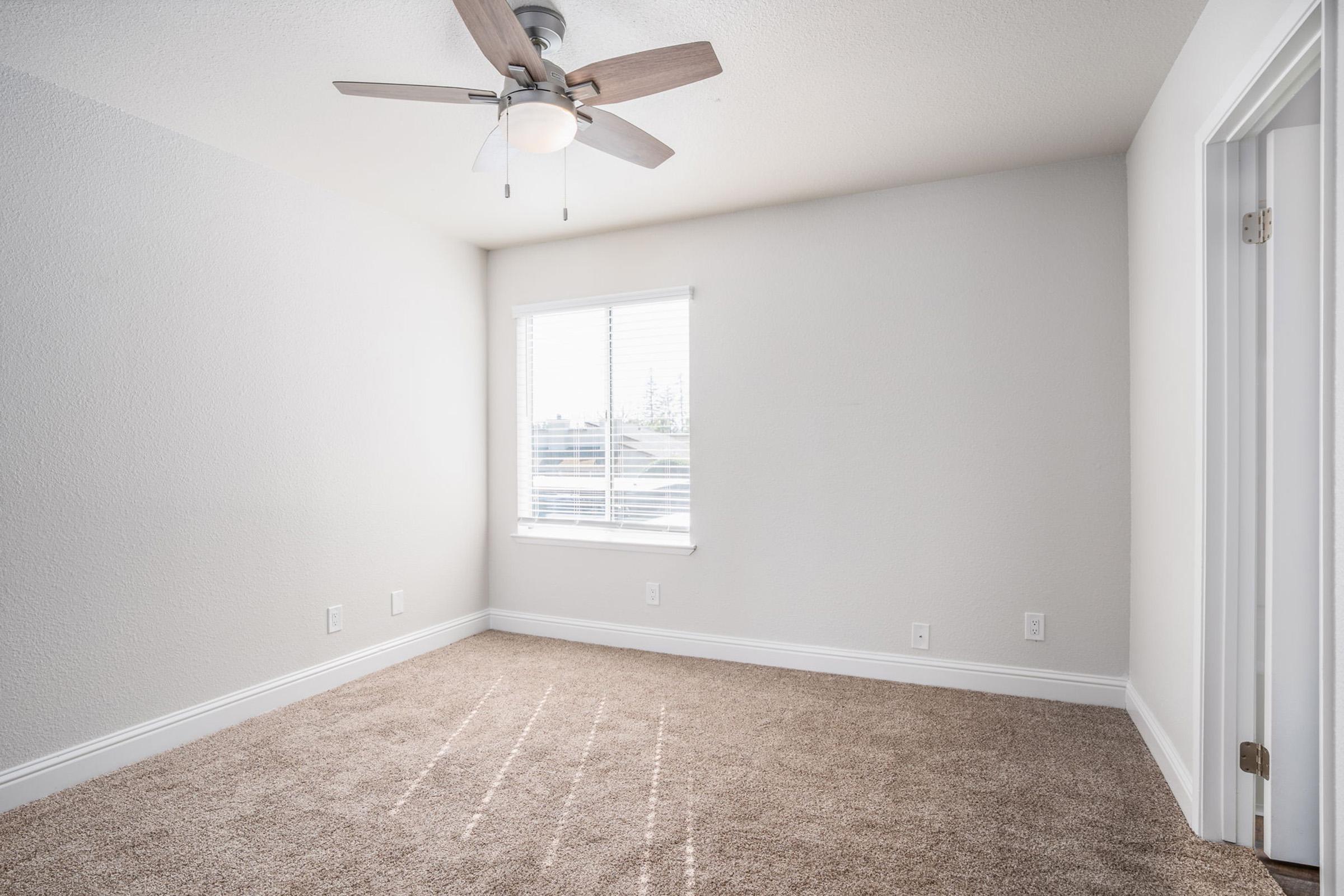 A bright and neutral bedroom with a ceiling fan, soft carpet, and a window letting in natural light. The walls are painted light gray, and there are two doors leading out, one likely a closet. The room is empty, creating a spacious feel.