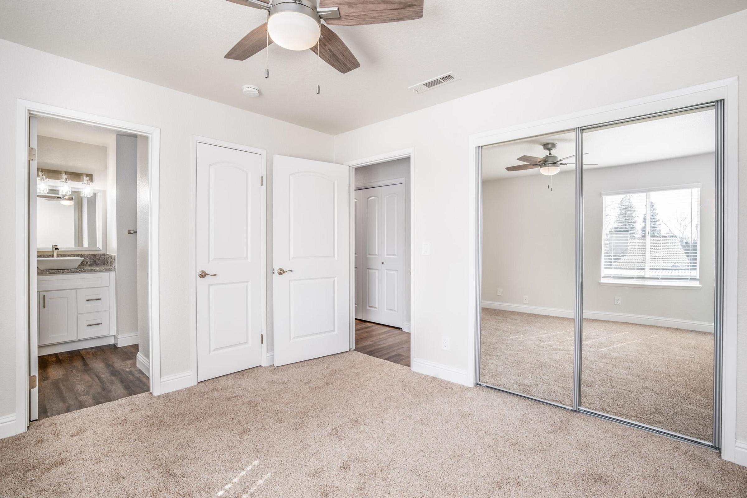 A spacious bedroom with beige carpet, white walls, and a ceiling fan. Two closed doors lead to other areas, and a mirrored closet door reflects the room. Natural light streams in through a window, highlighting a clean and modern aesthetic.