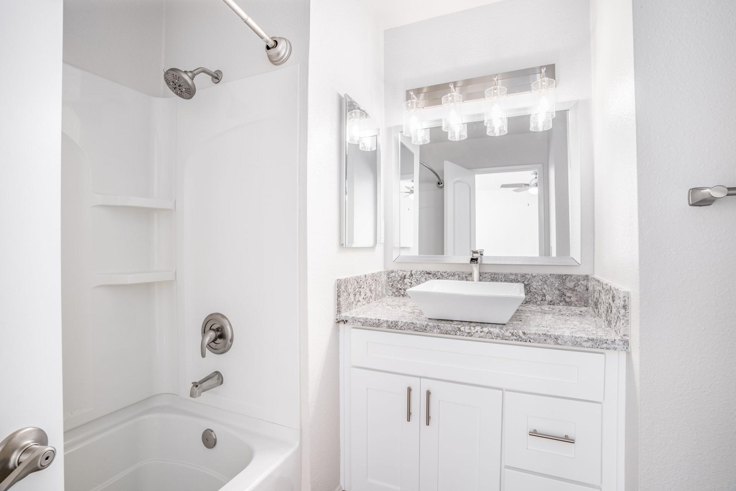 A modern bathroom featuring a bathtub with a showerhead, white tiled walls, and a sleek vanity. The vanity has a white vessel sink, granite countertop, and chrome fixtures. Bright lighting is provided by a row of glass pendant lights above the mirror, creating a clean and contemporary atmosphere.