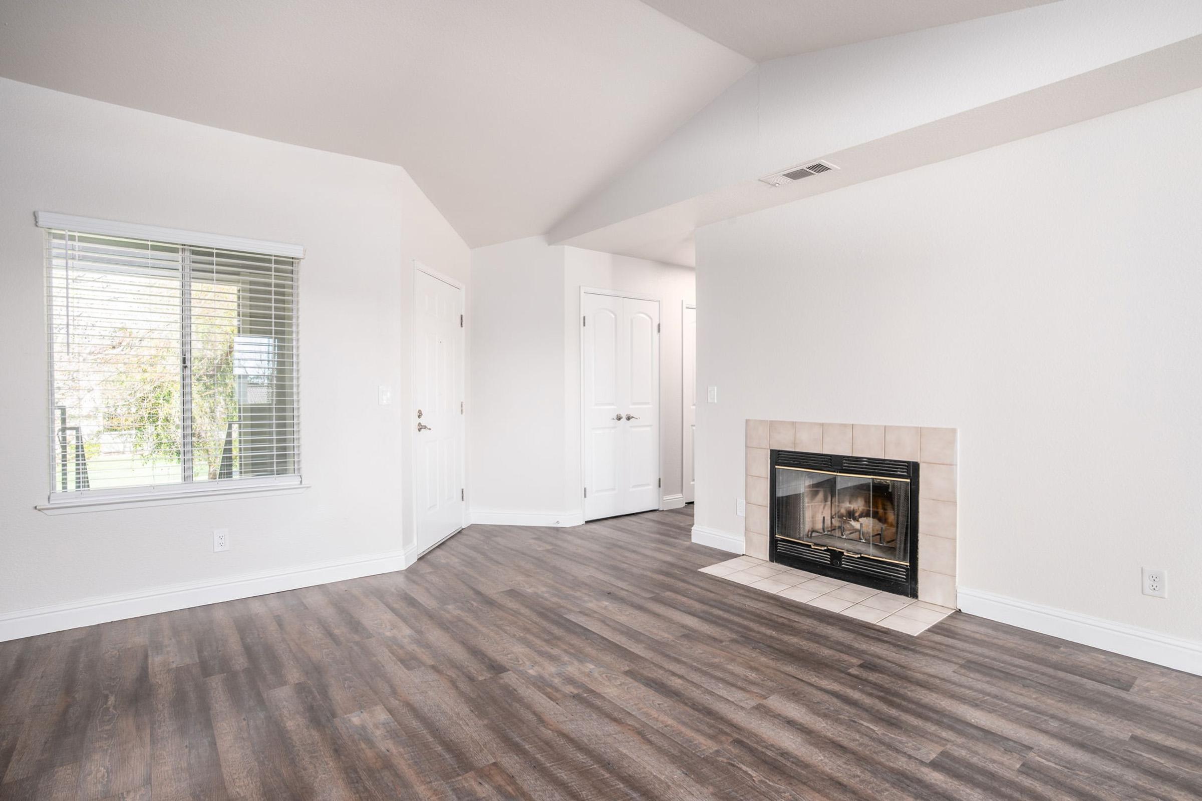 A spacious, empty living room featuring a hardwood floor, a fireplace with a tile surround, and large windows with white blinds. The walls are painted in light colors, and there are two doors leading to other areas of the home. The overall atmosphere is bright and airy.