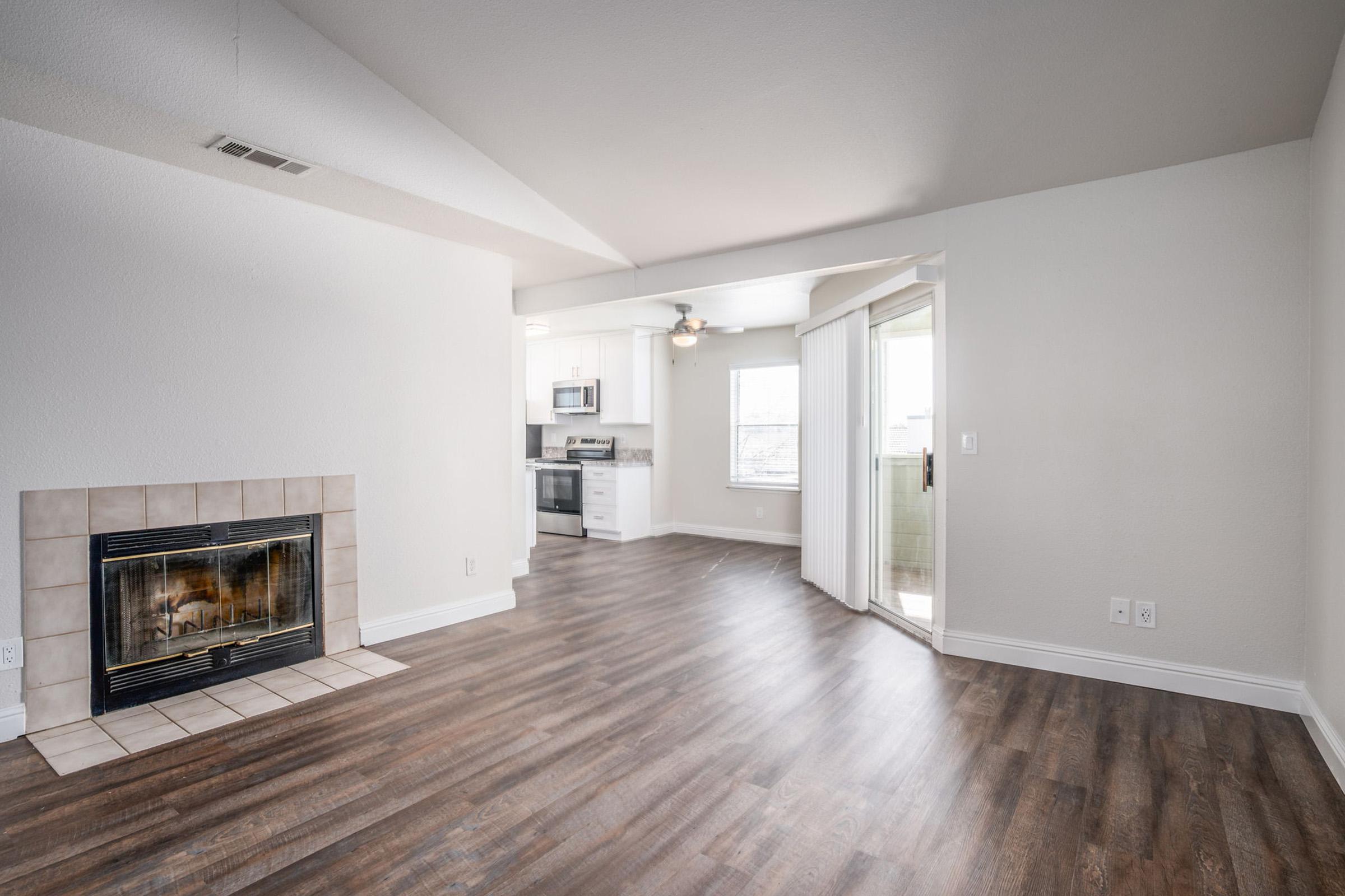 Interior of a modern living room featuring a fireplace, hardwood floors, and an open layout. The space includes a ceiling fan and leads to a kitchen with stainless steel appliances. Large windows allow natural light, and a door opens to a small balcony or porch area. The walls are painted in neutral colors.