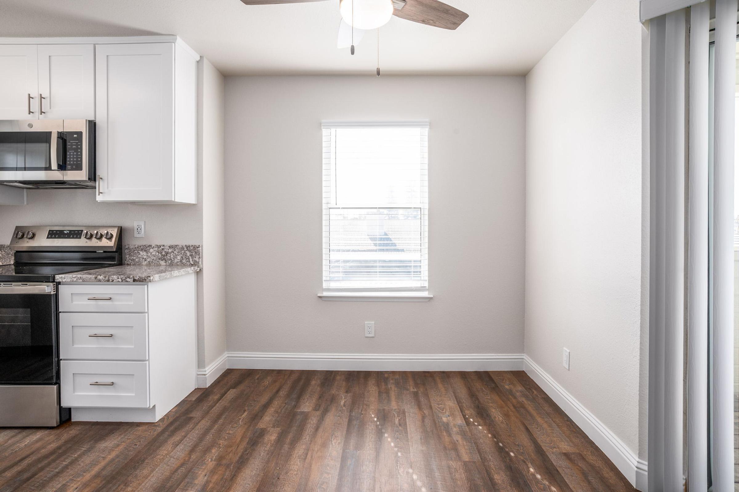 A modern kitchen space with a stainless steel stove, microwave, and white cabinetry. The room features a window with blinds, allowing natural light, and has light wood flooring that adds warmth to the area. A ceiling fan is visible, enhancing the airy atmosphere of the room.