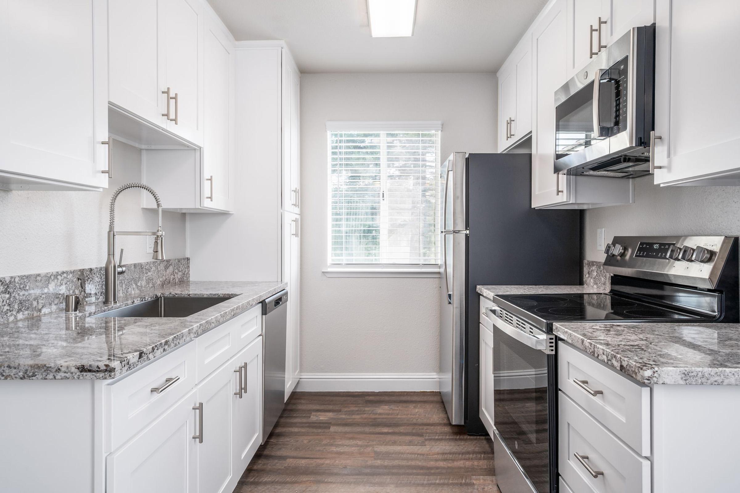 A modern kitchen featuring white cabinetry, granite countertops, a stainless steel sink with a pull-down faucet, a built-in microwave, an electric range, and a refrigerator. Natural light streams in through a window above the sink, highlighting the clean and sleek design of the space.