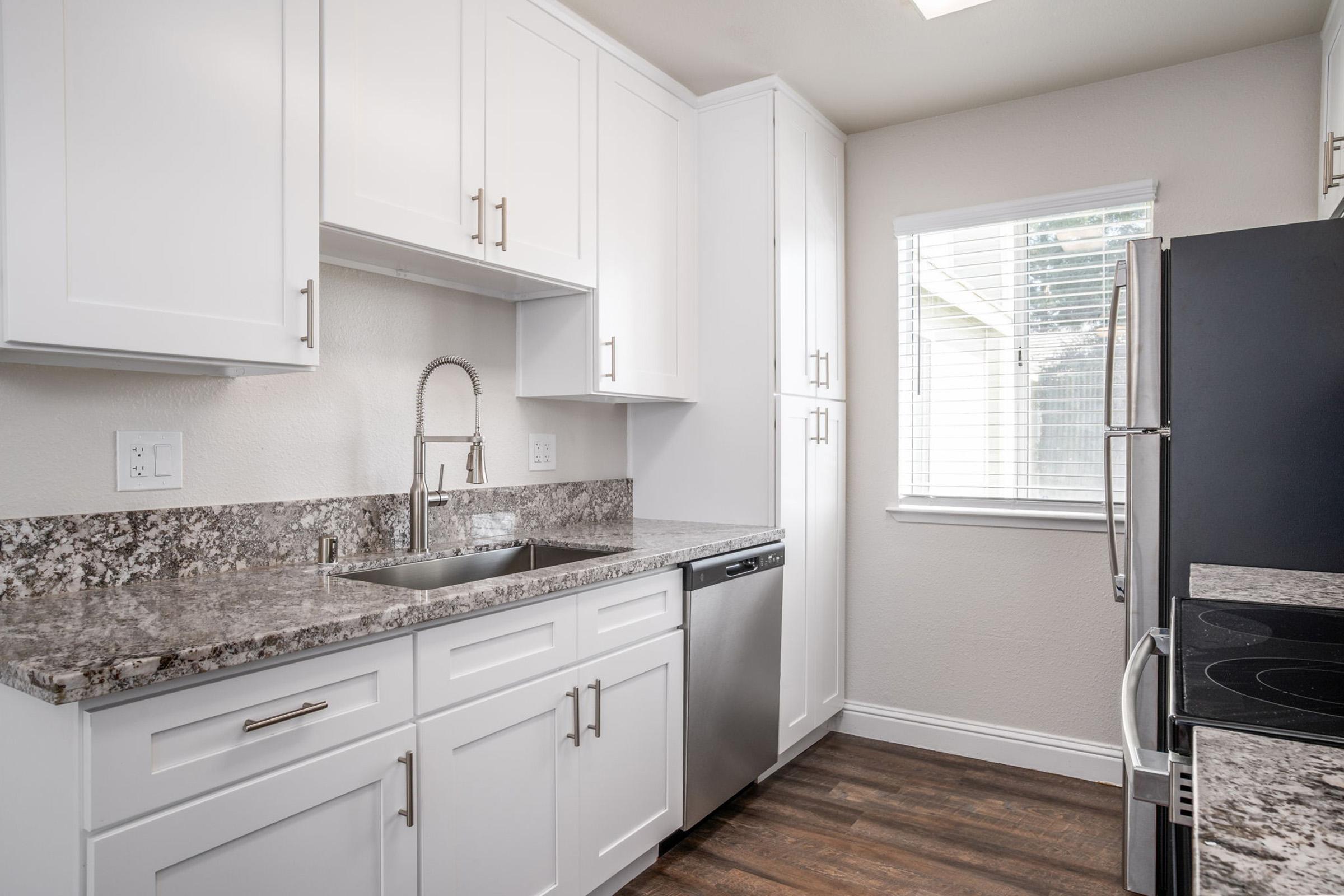 Modern kitchen featuring white cabinetry, a stainless steel sink with a faucet, and a dishwasher. The countertops are made of speckled granite. Appliances include a black refrigerator and a stovetop. Natural light enters through a window with blinds, illuminating the room. The flooring is dark wood.