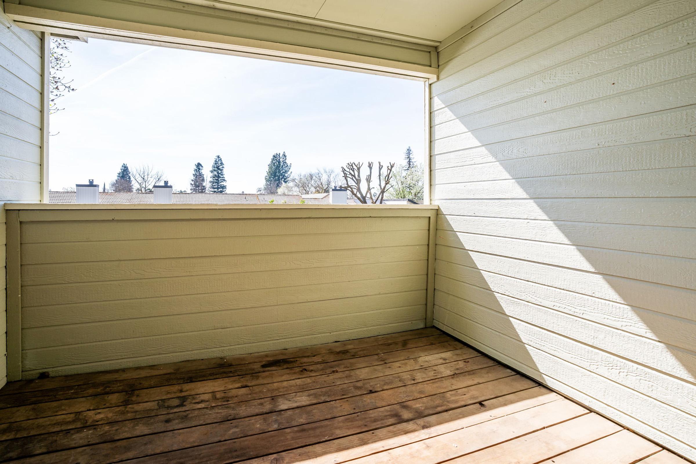 Empty balcony or porch with wooden flooring and light-colored walls. A large window provides a view of trees and distant rooftops. Sunlight casts shadows inside, creating a bright and airy atmosphere.