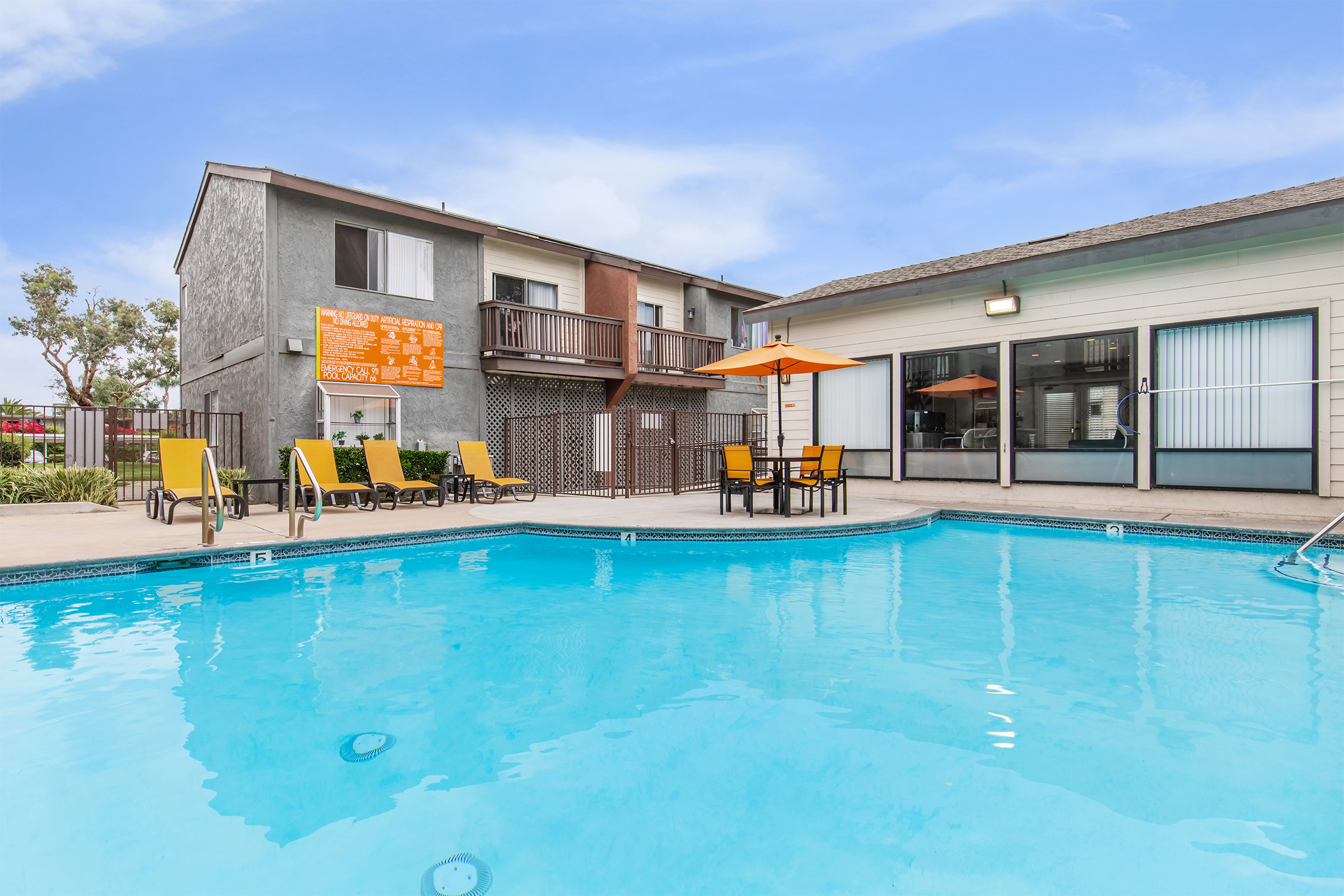 A clear blue swimming pool with yellow lounge chairs and an orange umbrella. In the background, there is a two-story building with a balcony and large windows. A dining table with chairs is set up near the pool area, creating a sunny and inviting outdoor atmosphere.