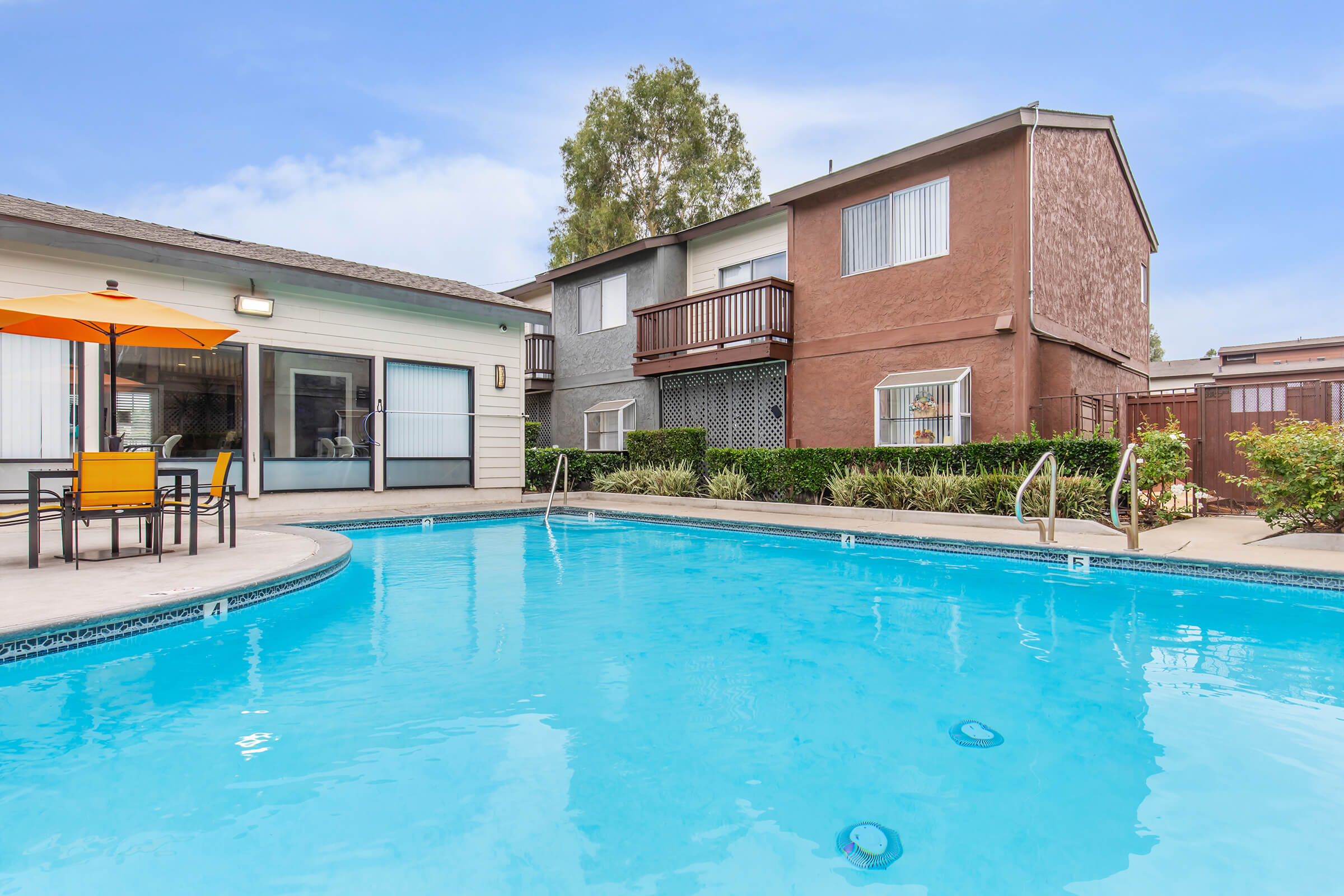 A sparkling blue swimming pool surrounded by a patio area with chairs and an umbrella. In the background, there are two multi-story apartment buildings with balconies, and lush greenery providing a serene atmosphere. The sky is clear and bright, adding to the inviting ambiance.