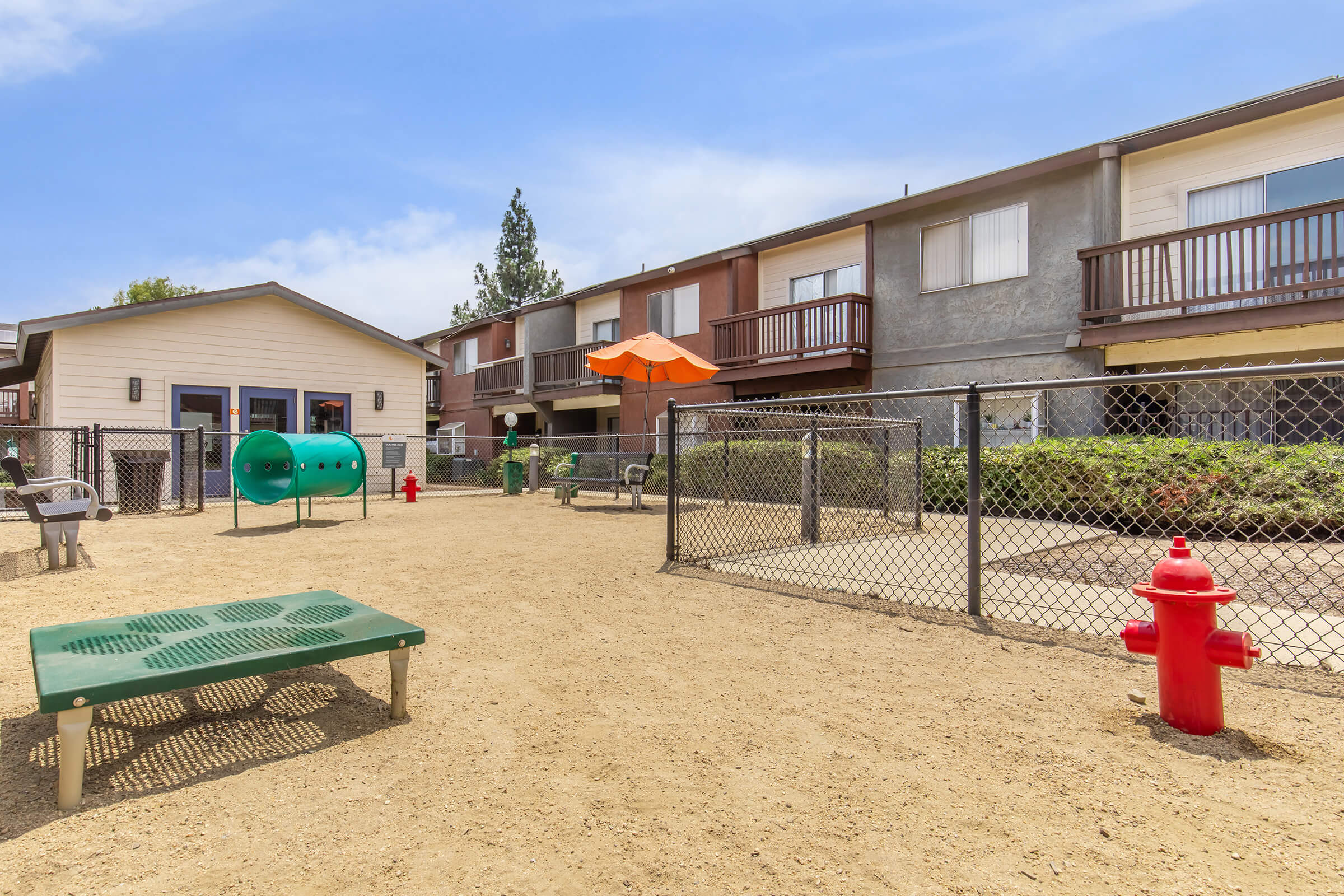 Playground area with sandy ground, featuring a green play structure, benches, and a red fire hydrant. There are fenced sections with ornamental plants, along with a building in the background that has a few balconies and an orange umbrella. Bright, sunny day with clear blue skies.