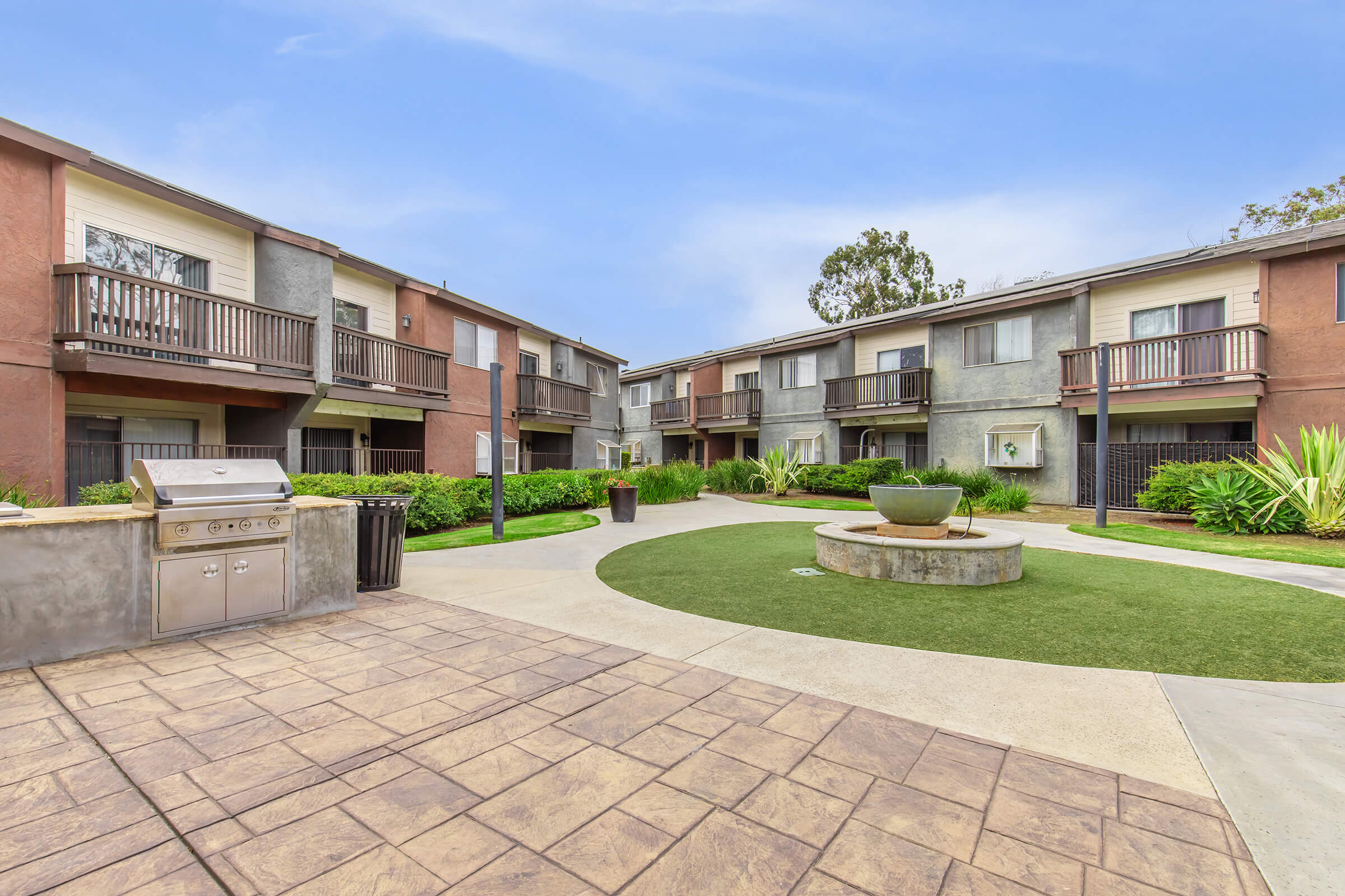 A landscaped courtyard with modern apartment buildings on either side. The pathway is paved with decorative concrete and leads to a circular stone planter with greenery. There are barbecue grills and lush foliage surrounding the area, creating a welcoming outdoor space.